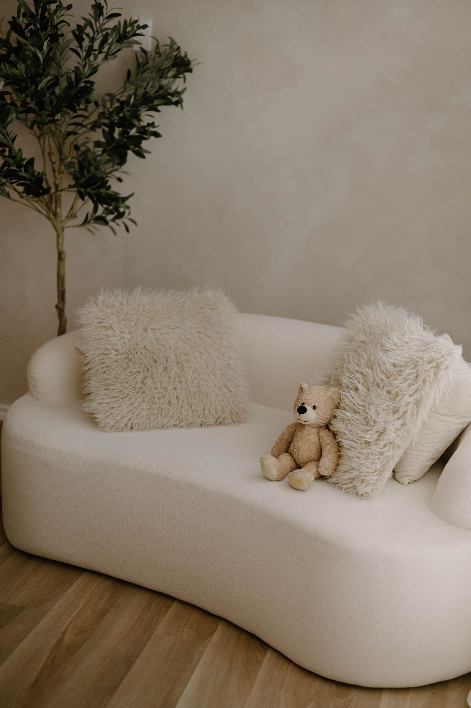 A white curved sofa with fluffy pillows and a teddy bear, beside a green plant on light wood flooring.