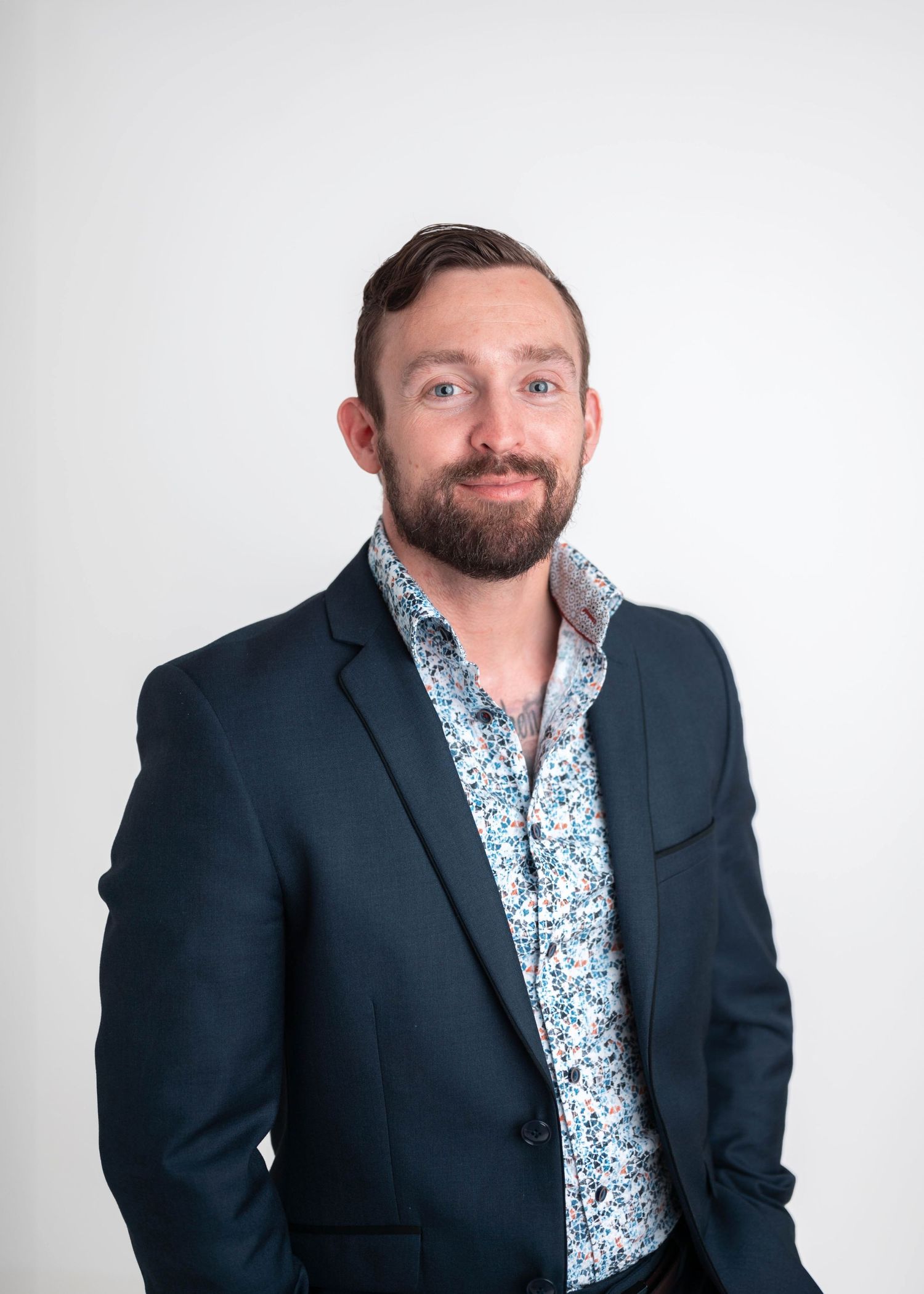 Professional man with beard wearing a floral shirt and dark blazer, smiling confidently against a white background.