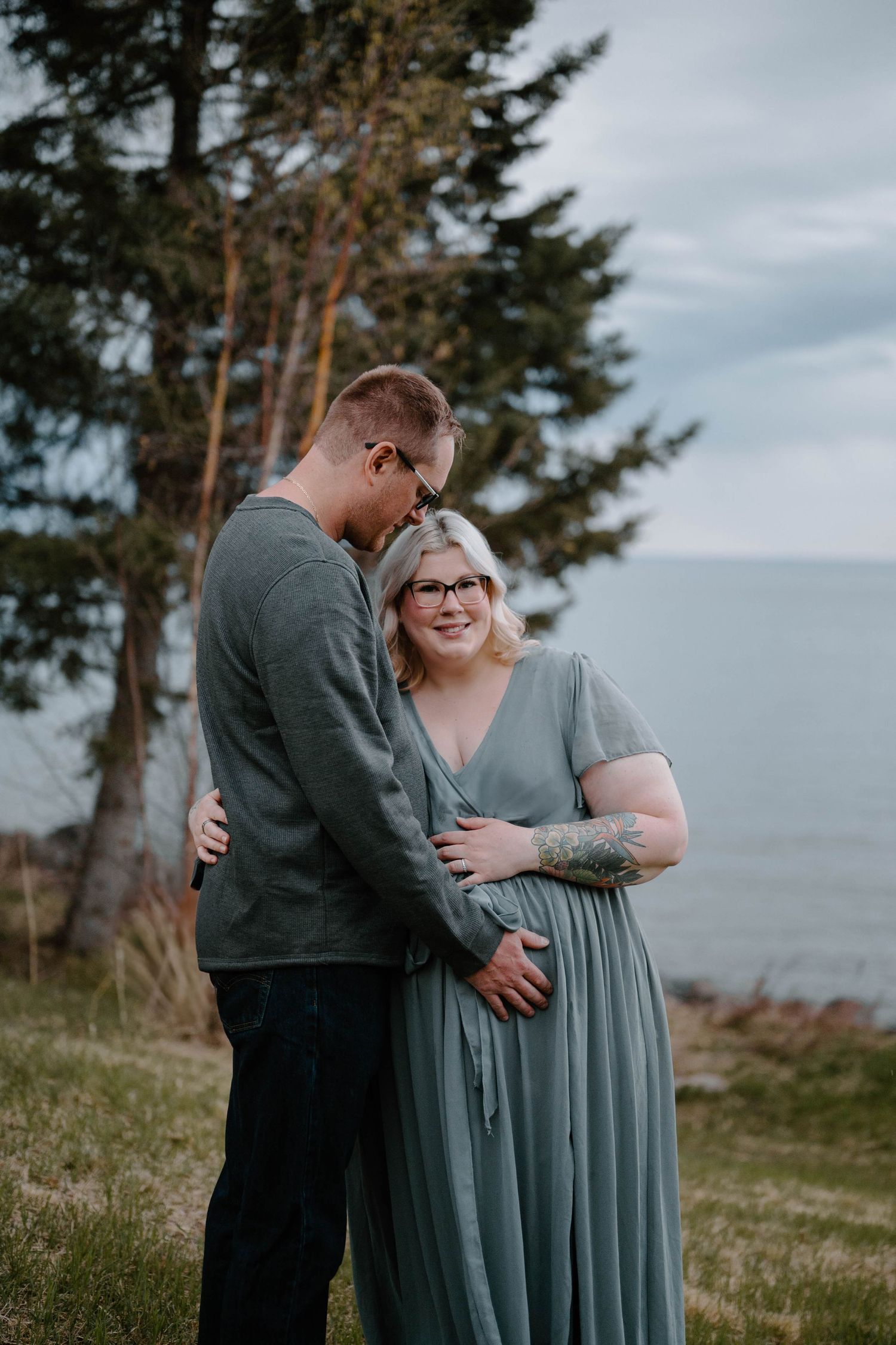 Couple in matching sage green outfits embracing outdoors near water, maternity photo session.