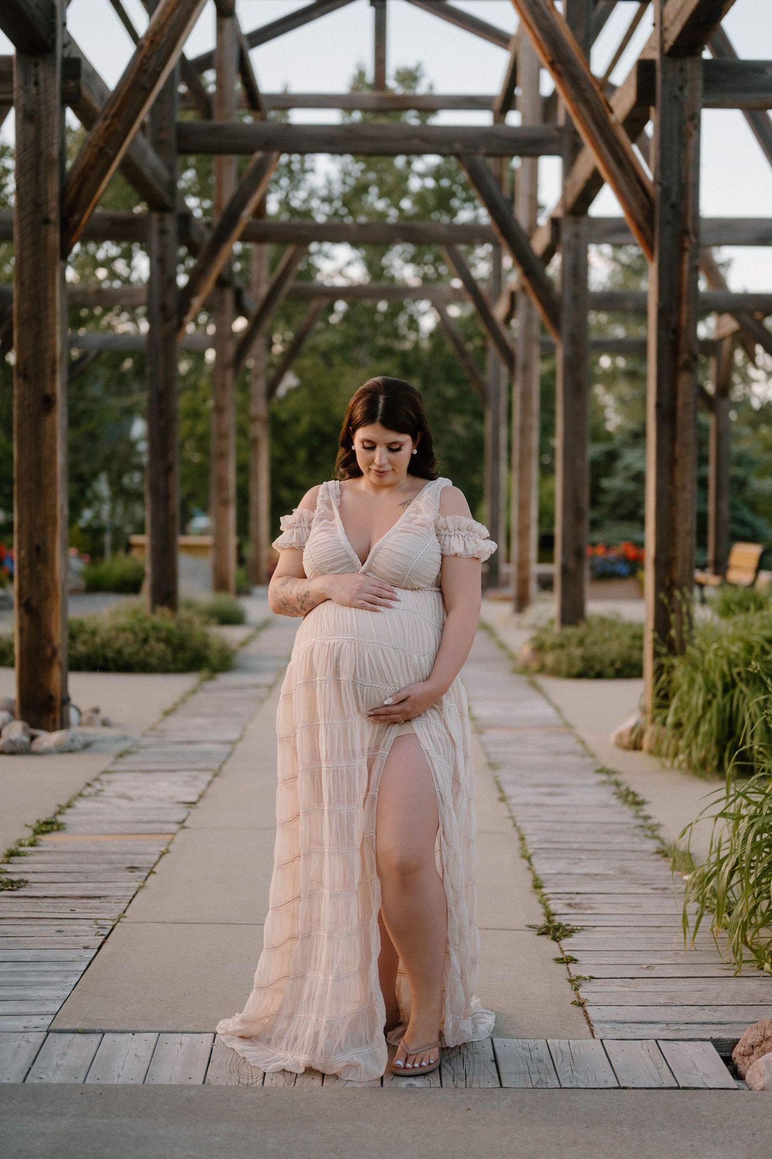 Pregnant woman in flowing blush gown cradling belly under rustic wooden pergola in a lush garden setting.