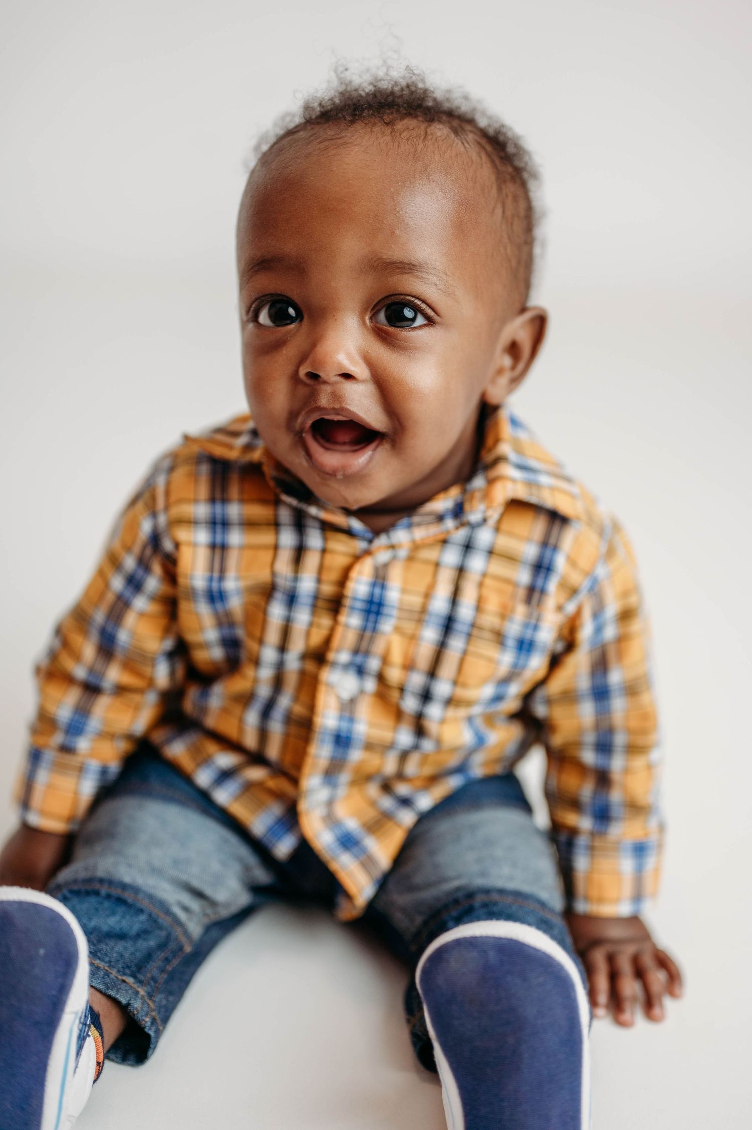 Adorable Black baby boy in a yellow plaid shirt and jeans sitting and laughing with mouth open.