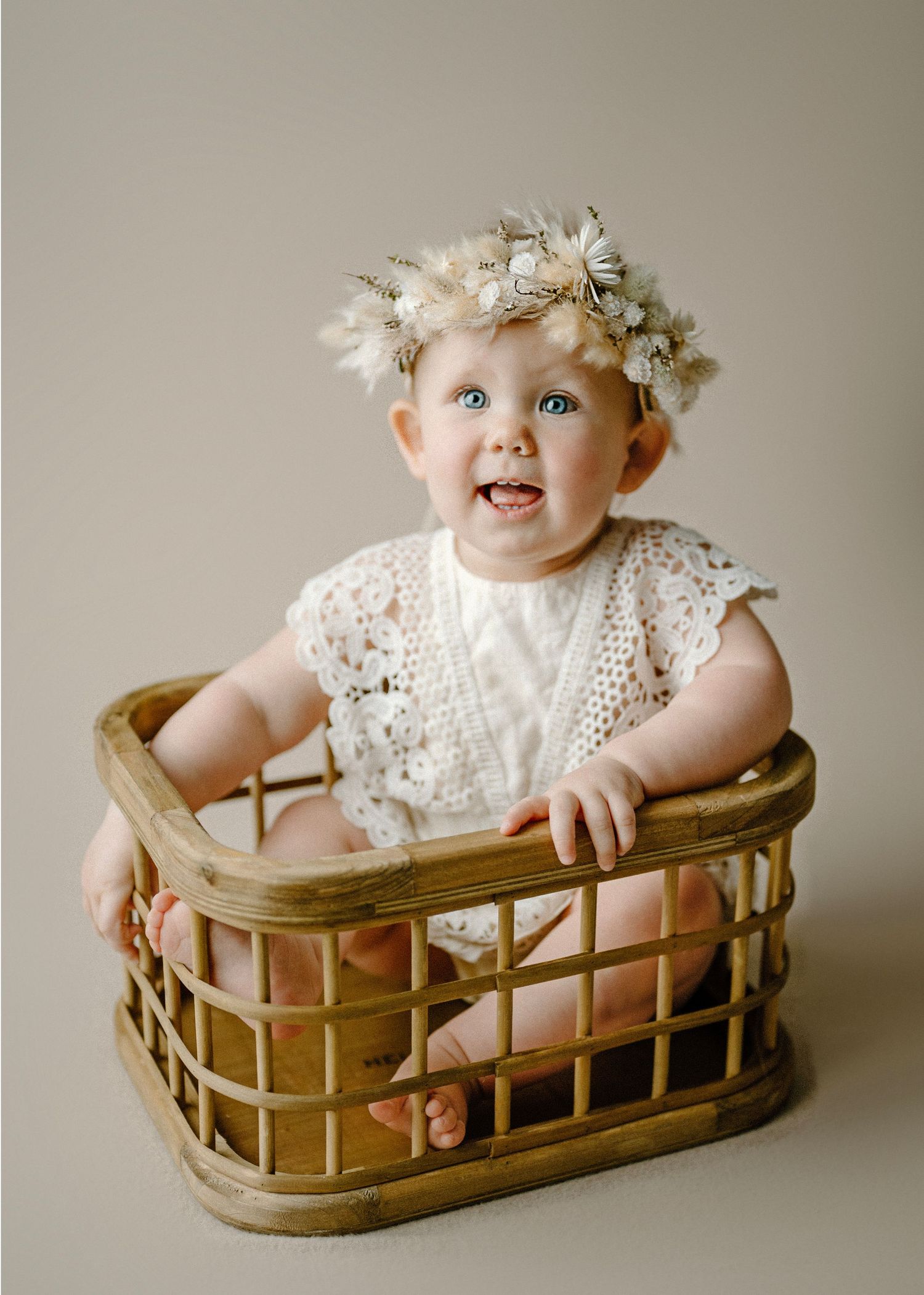 Happy blue-eyed baby wearing floral crown and white lace outfit sitting in wicker basket.