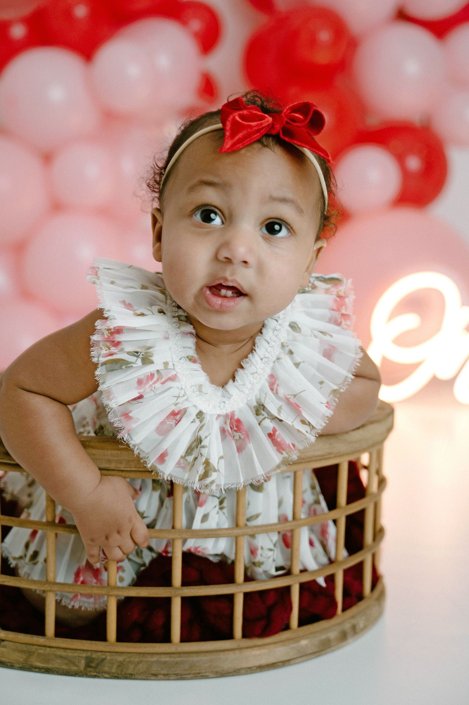 Close-up of baby girl in ruffled floral dress sitting in wicker basket with balloon background.