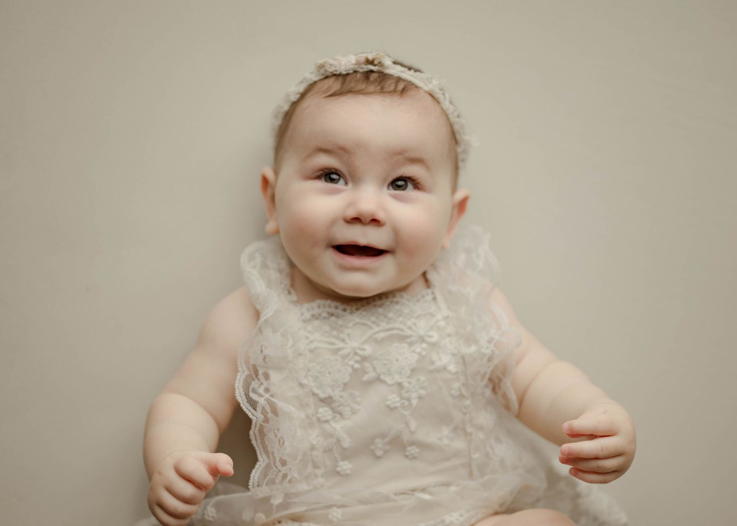 Smiling baby girl in white lace dress and headband lying down during professional studio portrait photography session.