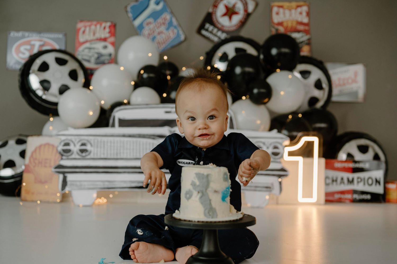 Baby boy in navy romper smashes first birthday cake at garage-themed photoshoot with number one sign.