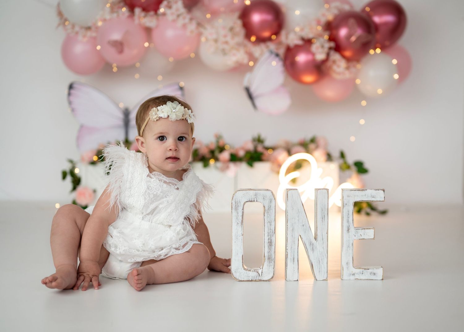 Baby girl in white outfit at floral butterfly first birthday cake smash with pink balloons.