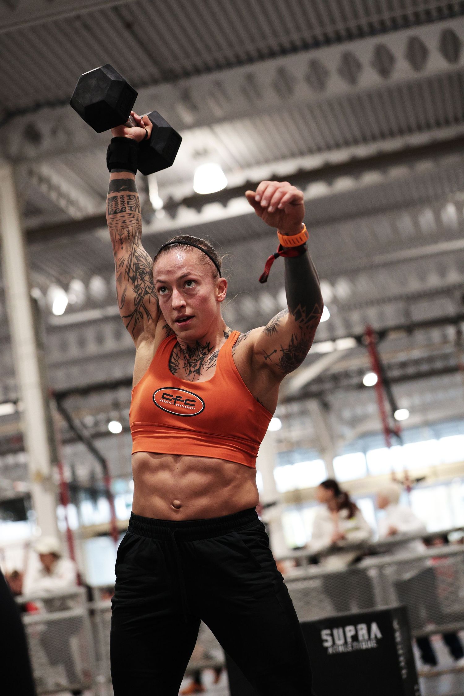Tattooed female athlete in orange crop top lifts dumbbell overhead during CrossFit competition in large gym.