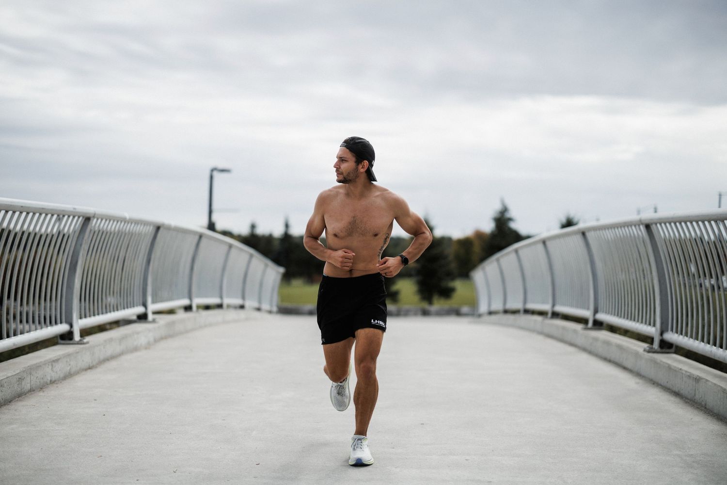 A shirtless runner jogs across a curved white bridge with metal railings on an overcast day.