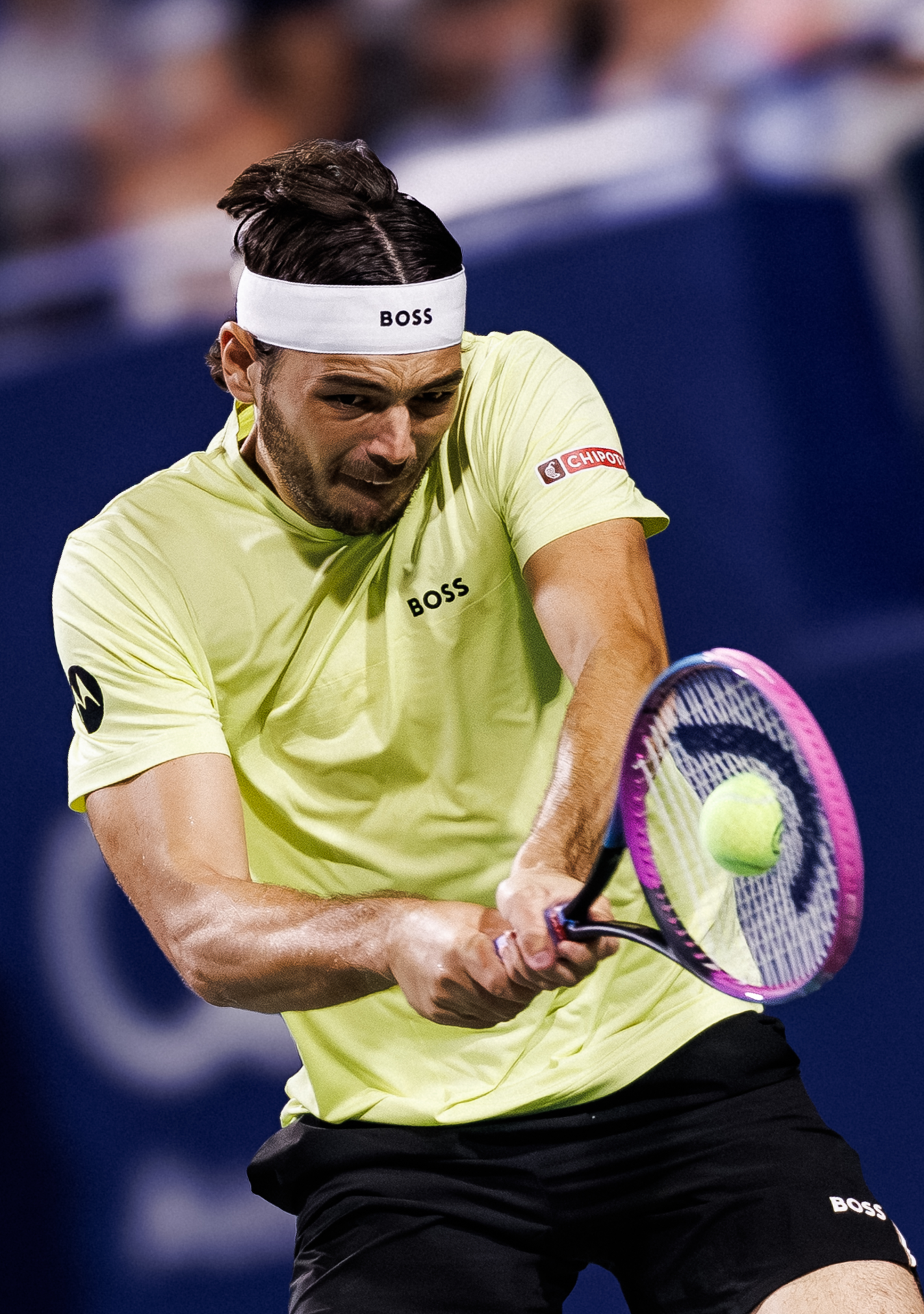 Tennis player wearing yellow shirt performs a backhand shot during a match on a dark court.