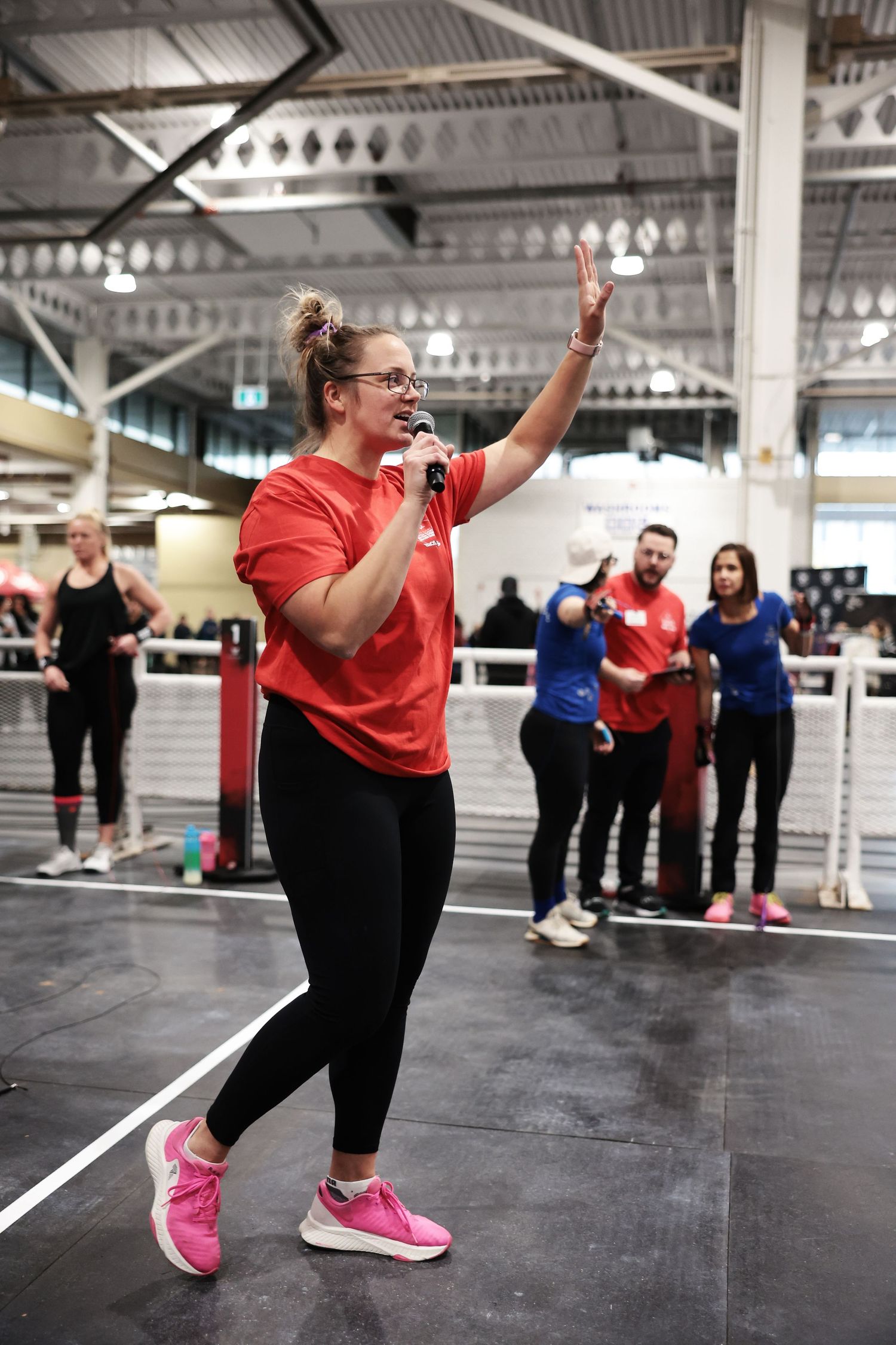 Woman in red t-shirt and pink sneakers holds microphone while leading a fitness class in a large indoor gym.