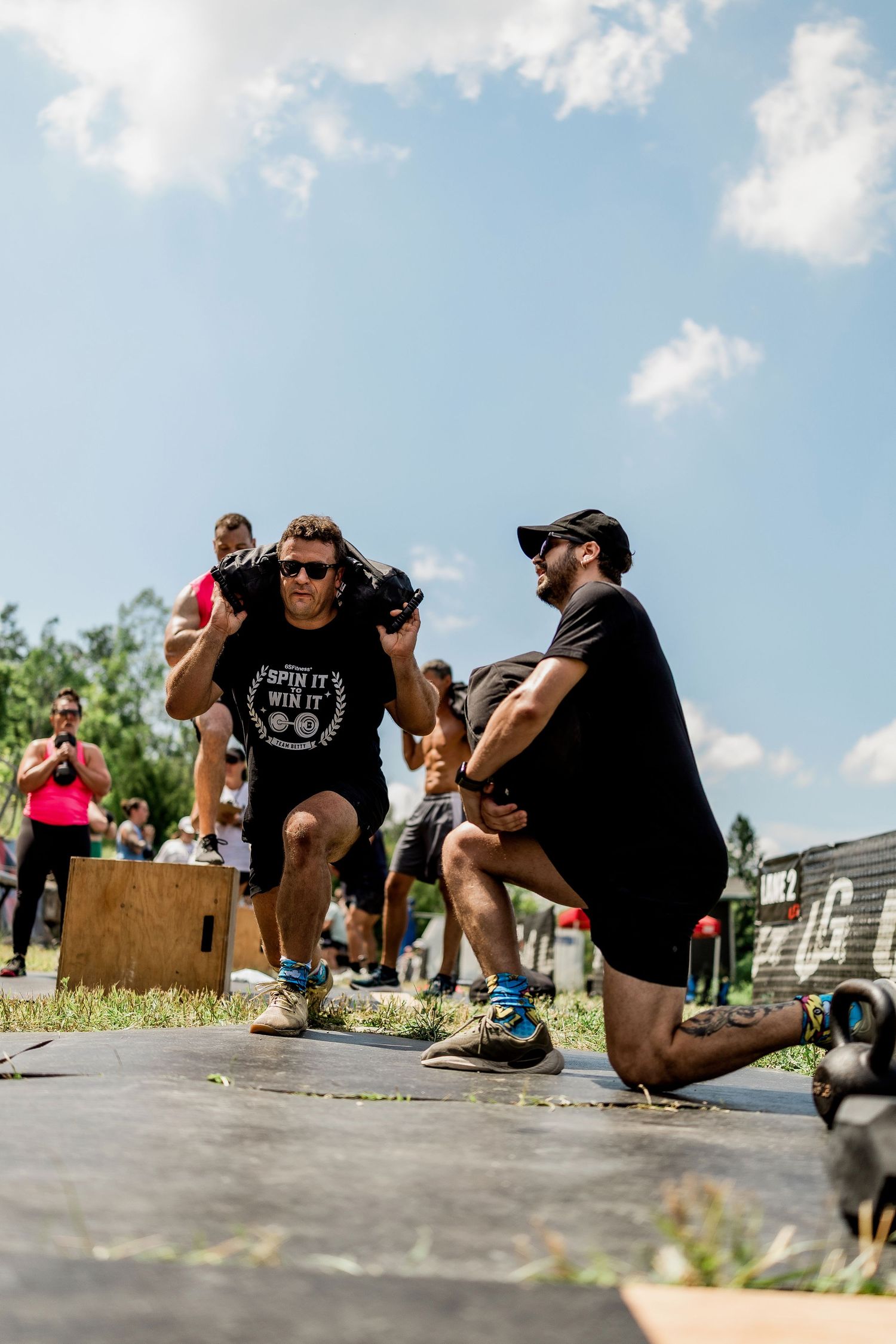 Two men doing lunges outdoors at a fitness competition on a sunny day, wearing black shirts and sunglasses.