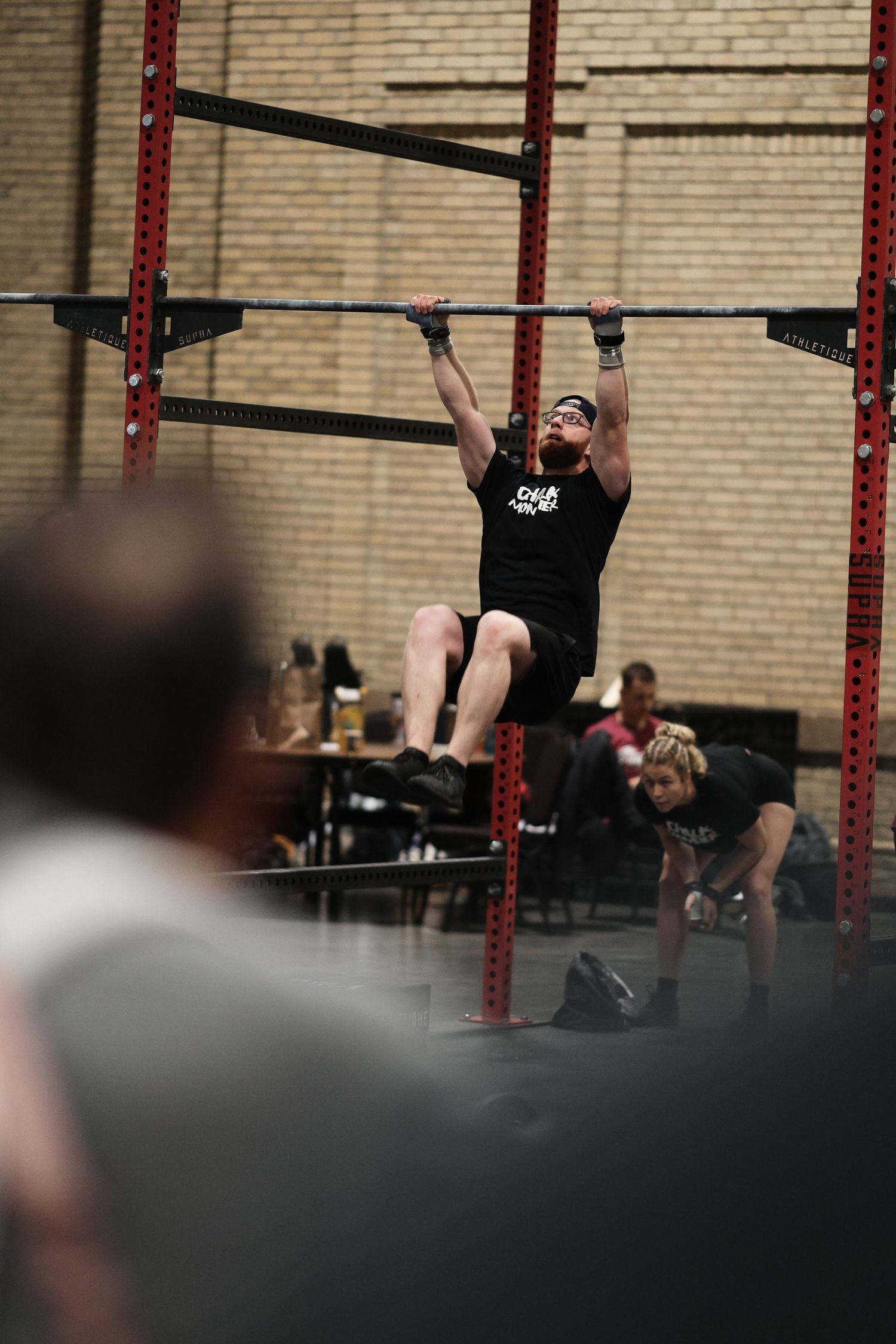 Athletes perform pull-ups and exercises on a rig against a brick wall in a CrossFit-style gym setting.