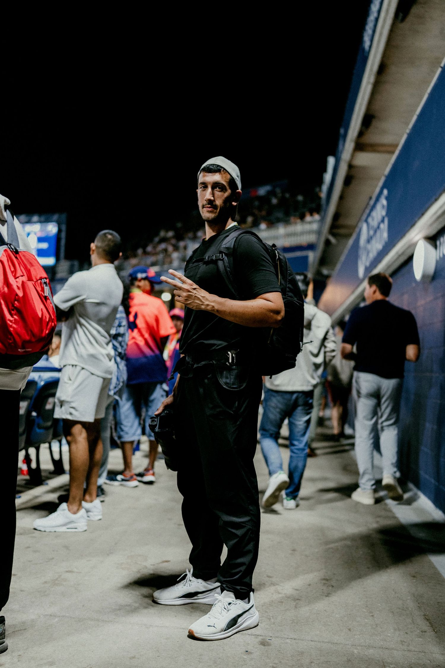 Casual spectator in black outfit and beanie standing in crowded stadium concourse area.