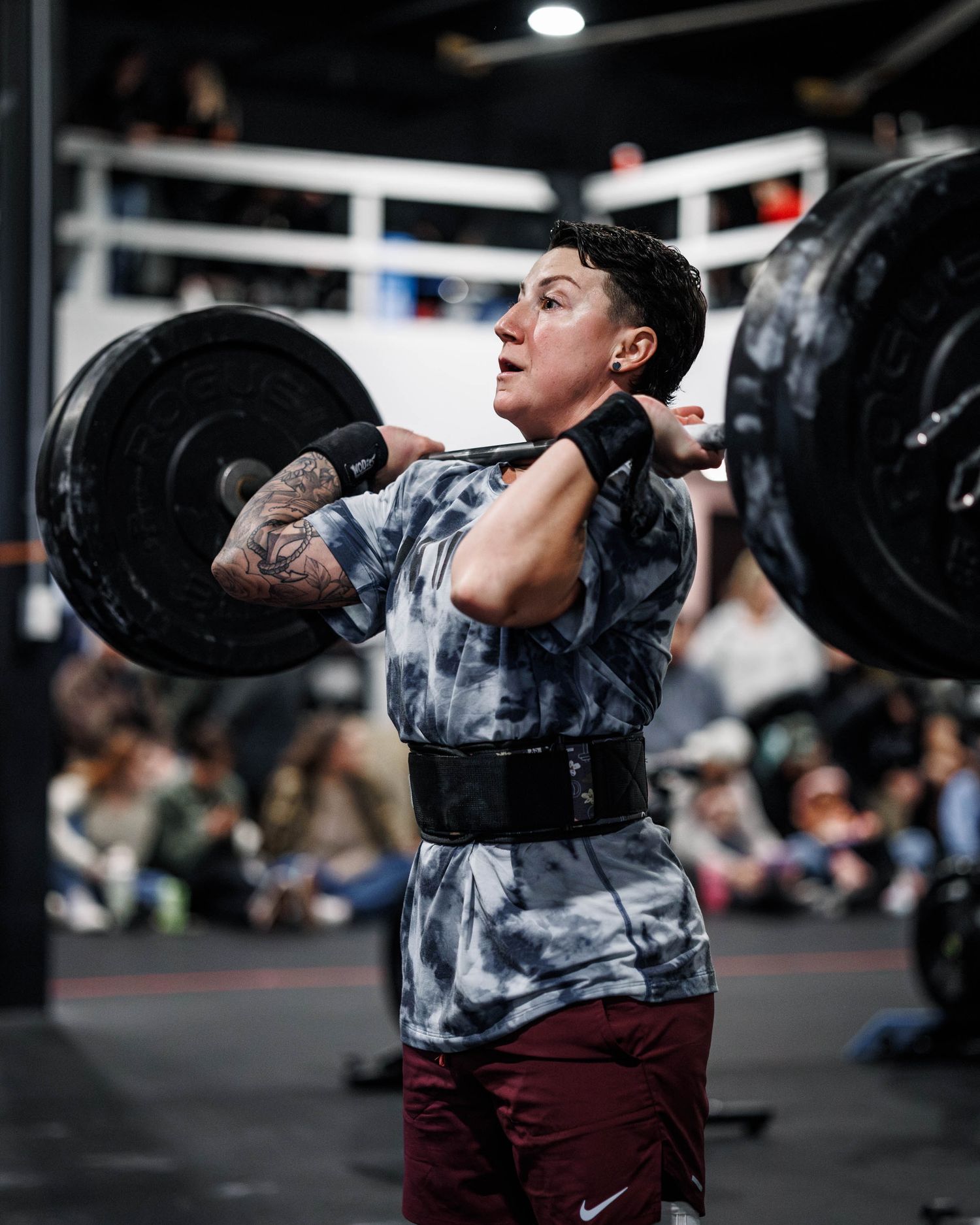 Weightlifter in tie-dye patterned shirt and maroon pants performs clean and jerk lift with barbell in competitive setting.