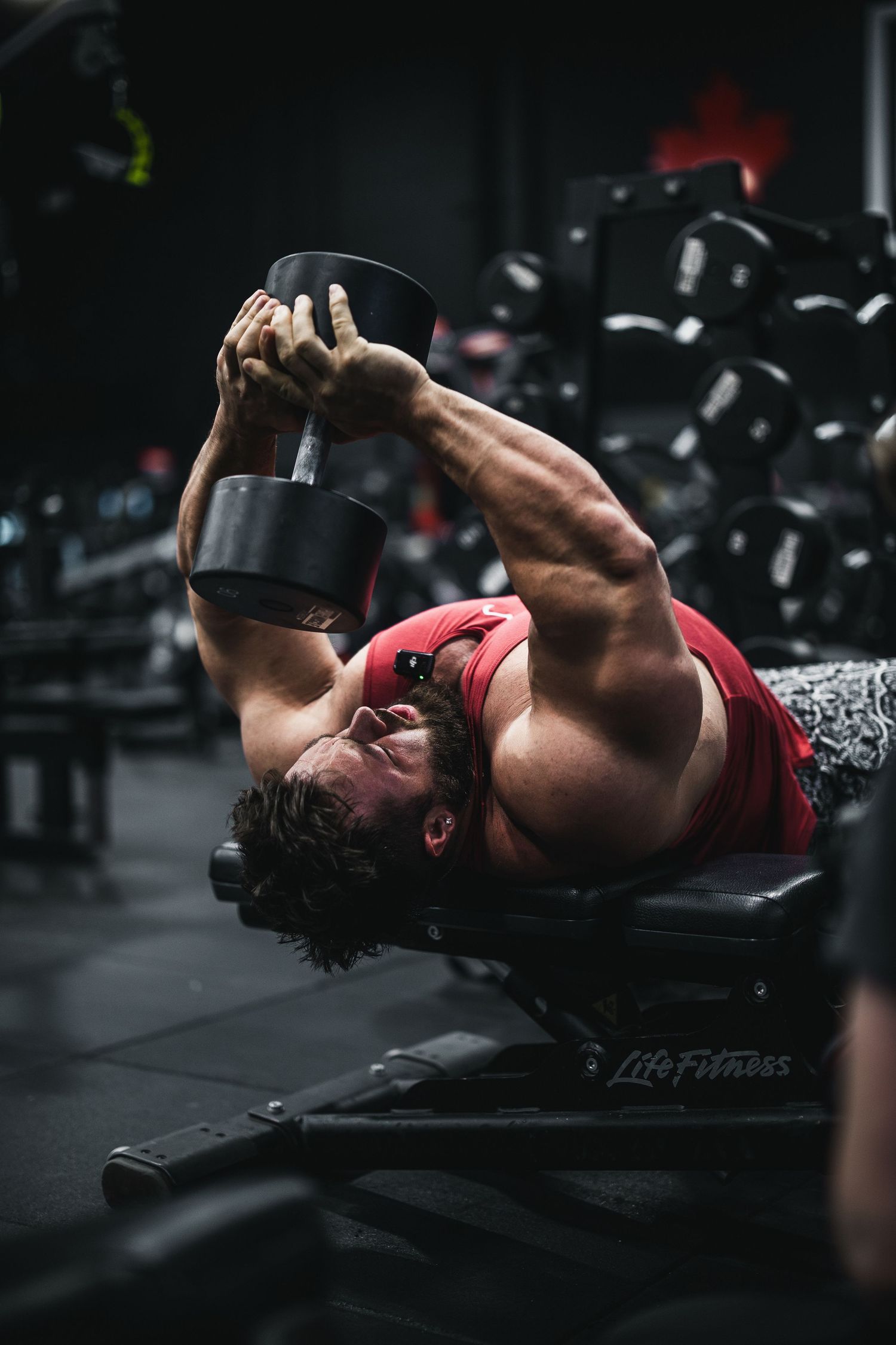 A person performs dumbbell chest presses lying on a bench in a dark gym setting with dramatic lighting.