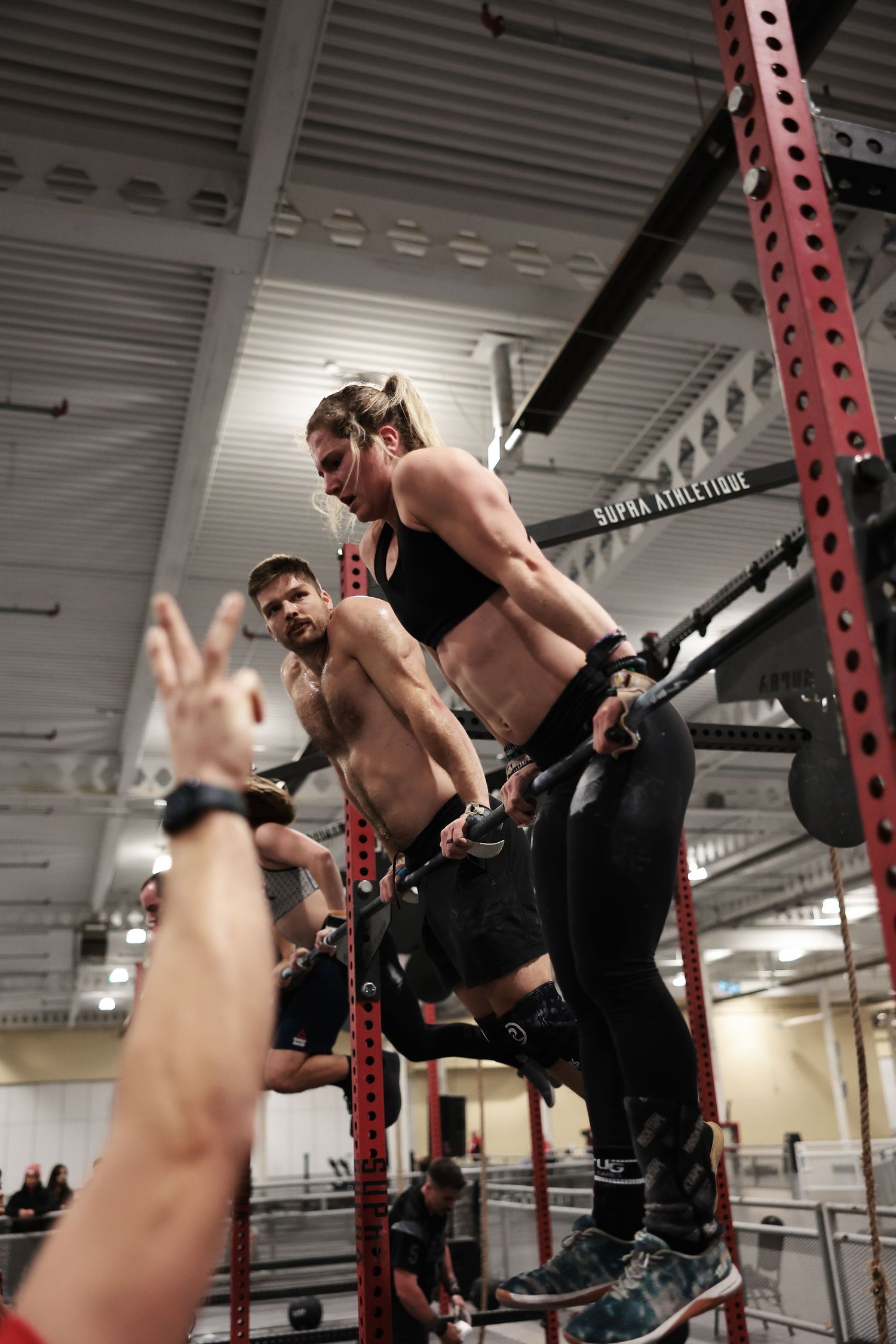 Athletic woman performing pull-ups on a red rack at Supra Athletique gym with a shirtless man cheering behind her.