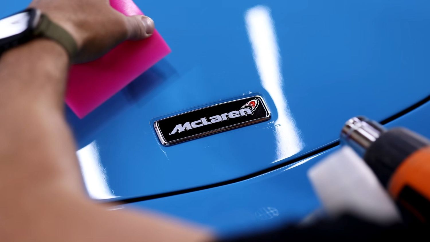 A car detailer applies vinyl decal protection film over a McLaren badge on a blue car.