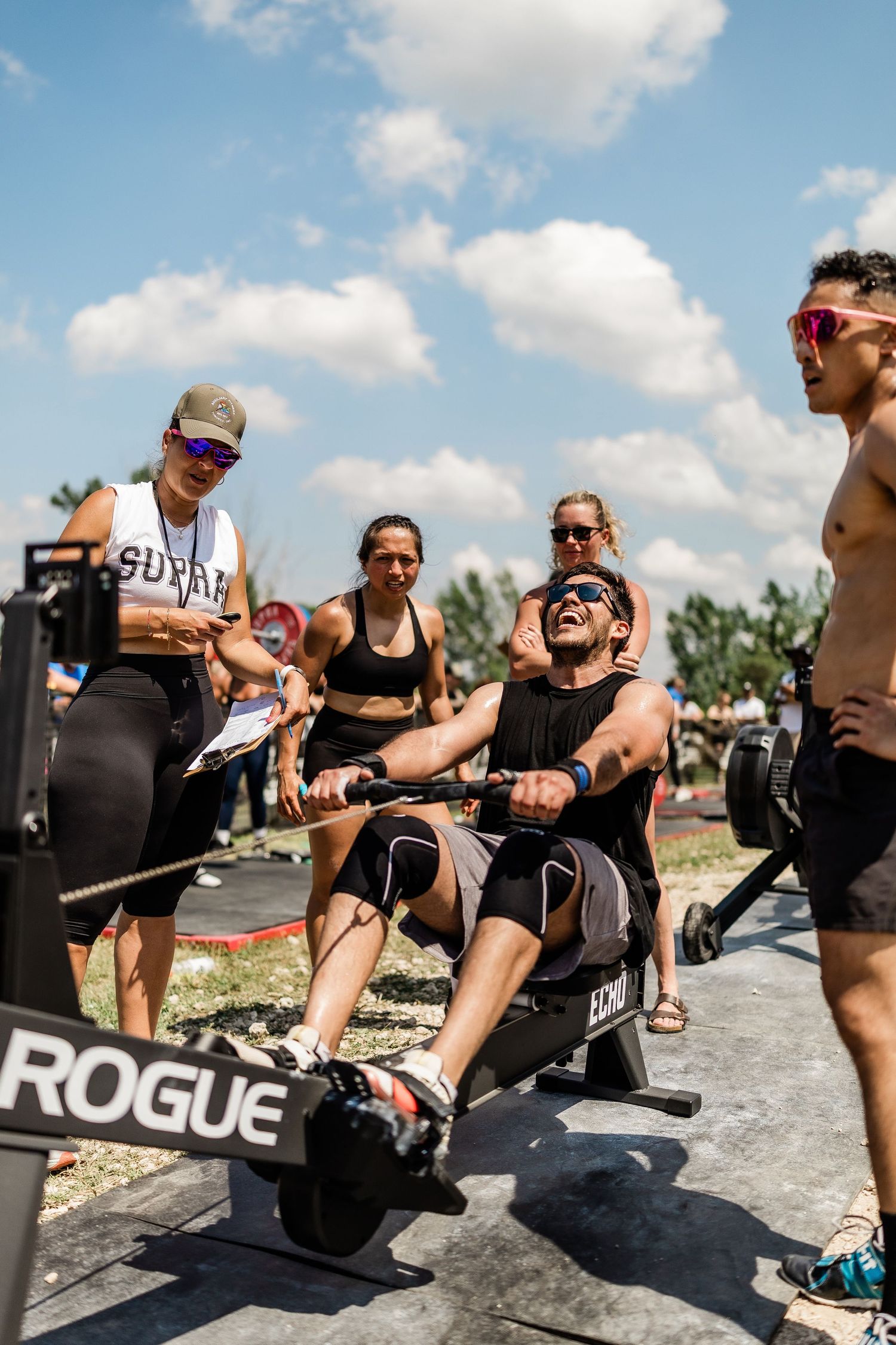 Athletes compete in outdoor CrossFit rowing event on Rogue machines, cheered on by spectators under a cloudy sky.