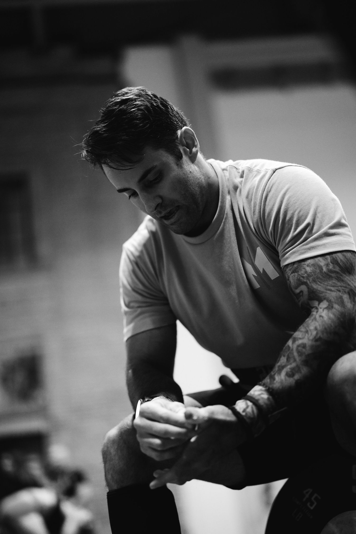 Black and white photo of an athlete in a white t-shirt with tattoos sitting and looking down during a workout.
