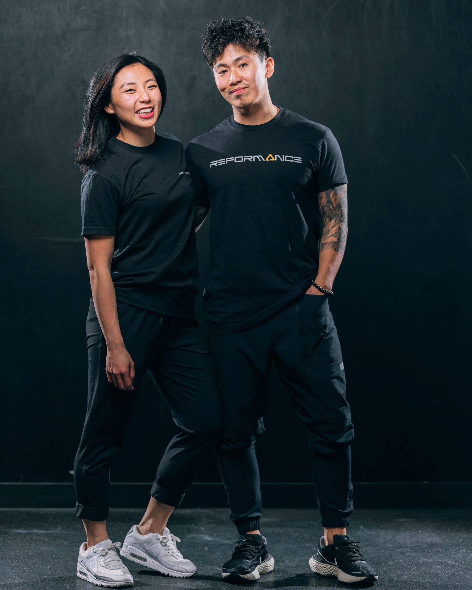 Two people in black athletic wear and sneakers pose together against a dark background for a fitness photoshoot.