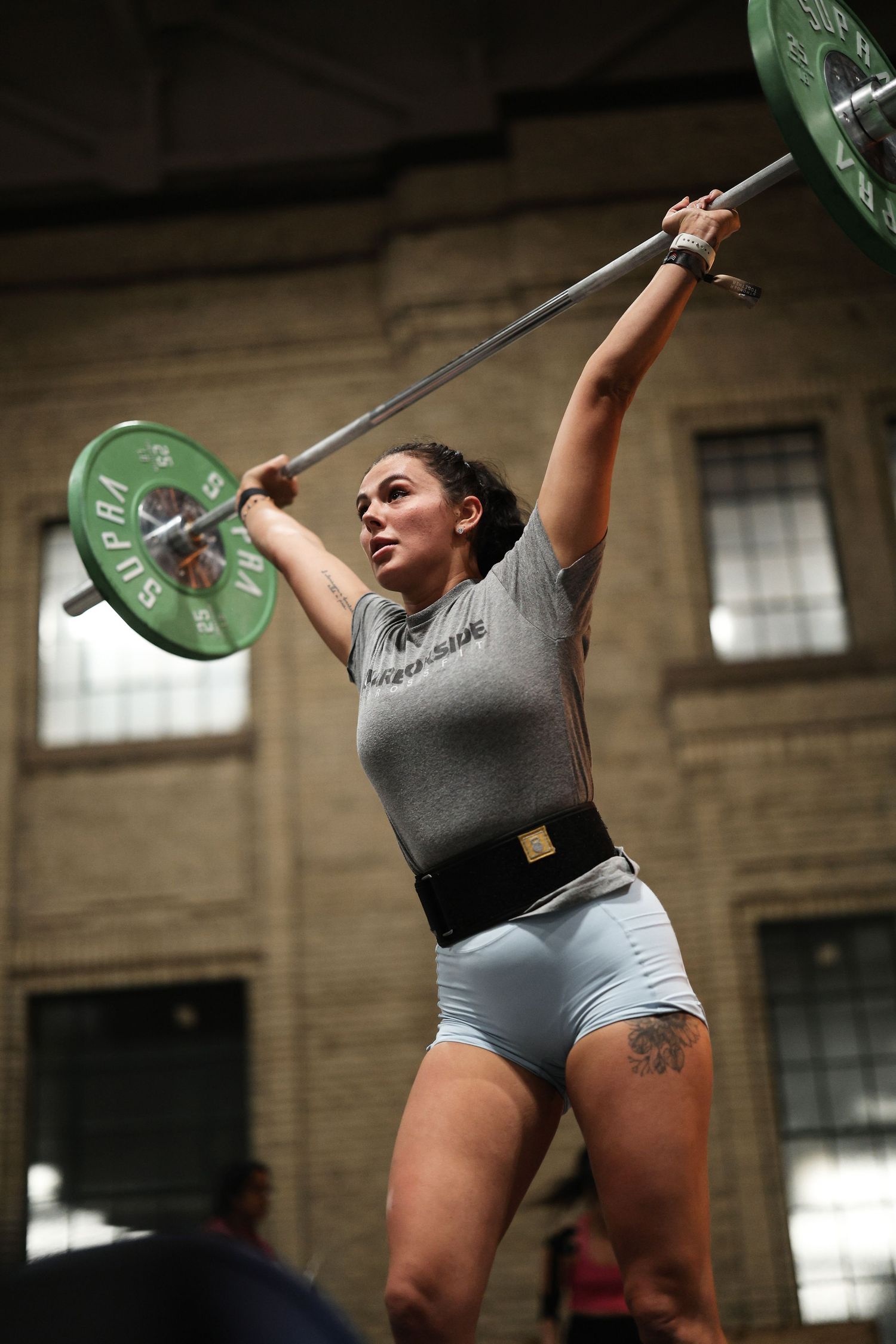 An athlete executes a barbell snatch lift in a warehouse-style gym setting with green plates.