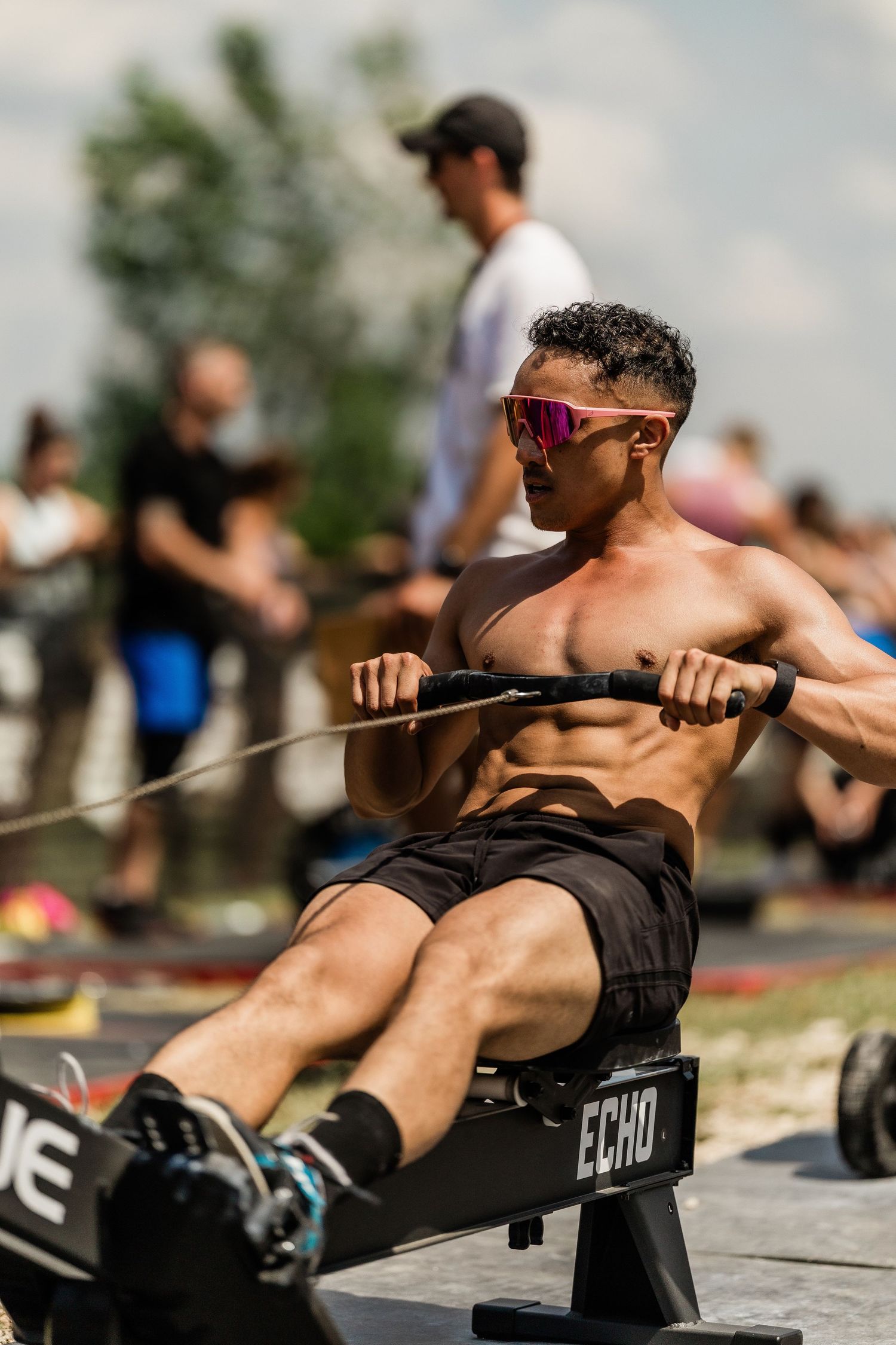 Muscular athlete in sunglasses performs rowing exercise on exercise machine during outdoor workout competition.