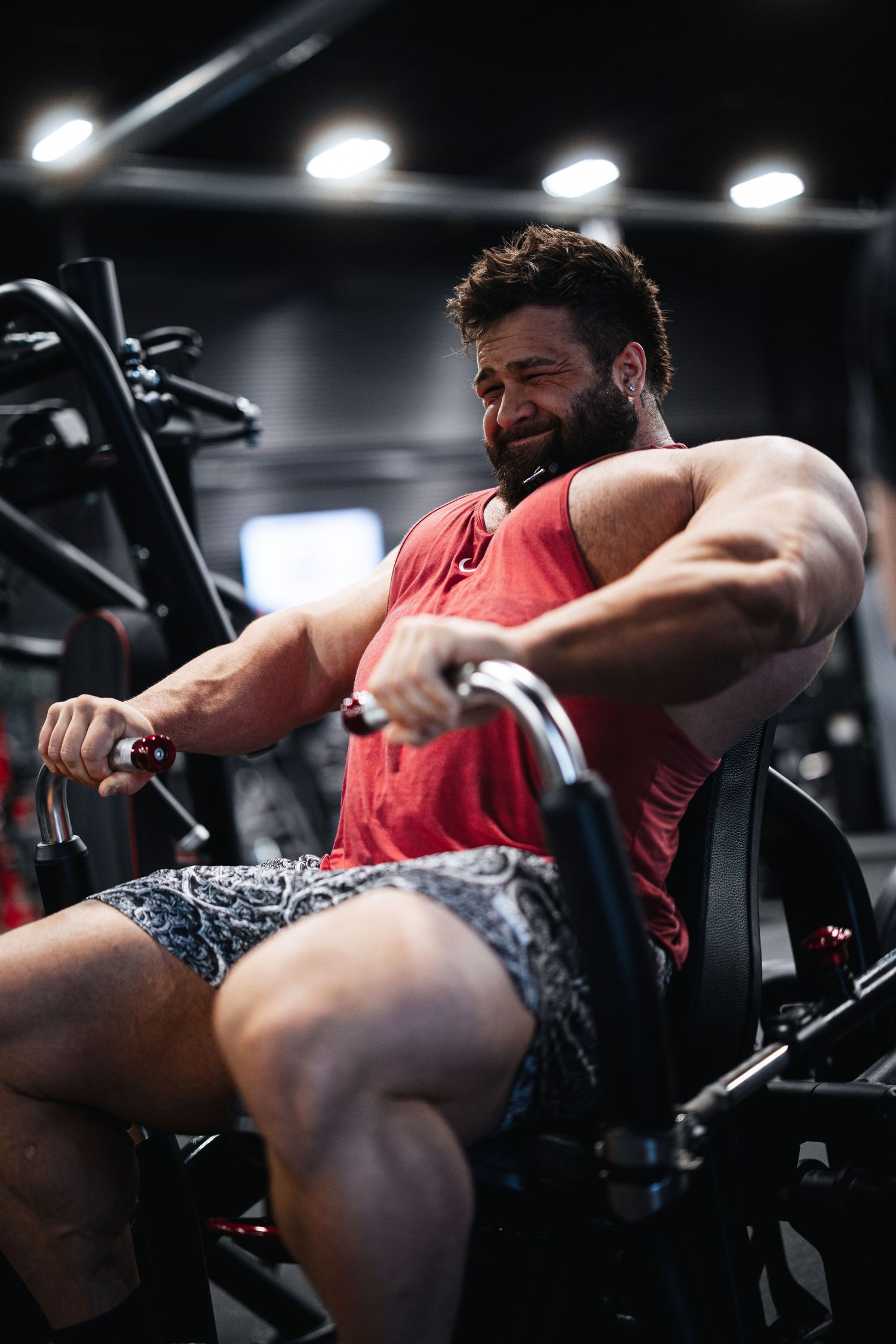 Muscular person wearing a red tank top performing an intense workout on a rowing machine in a dark gym.