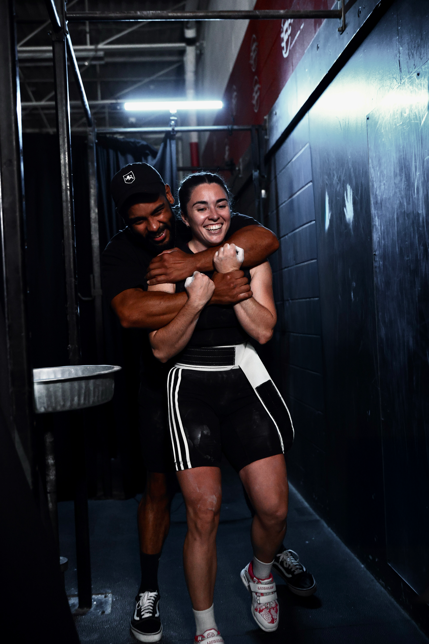 Two people playfully embrace and laugh together in a dark hallway with black walls and dim lighting.