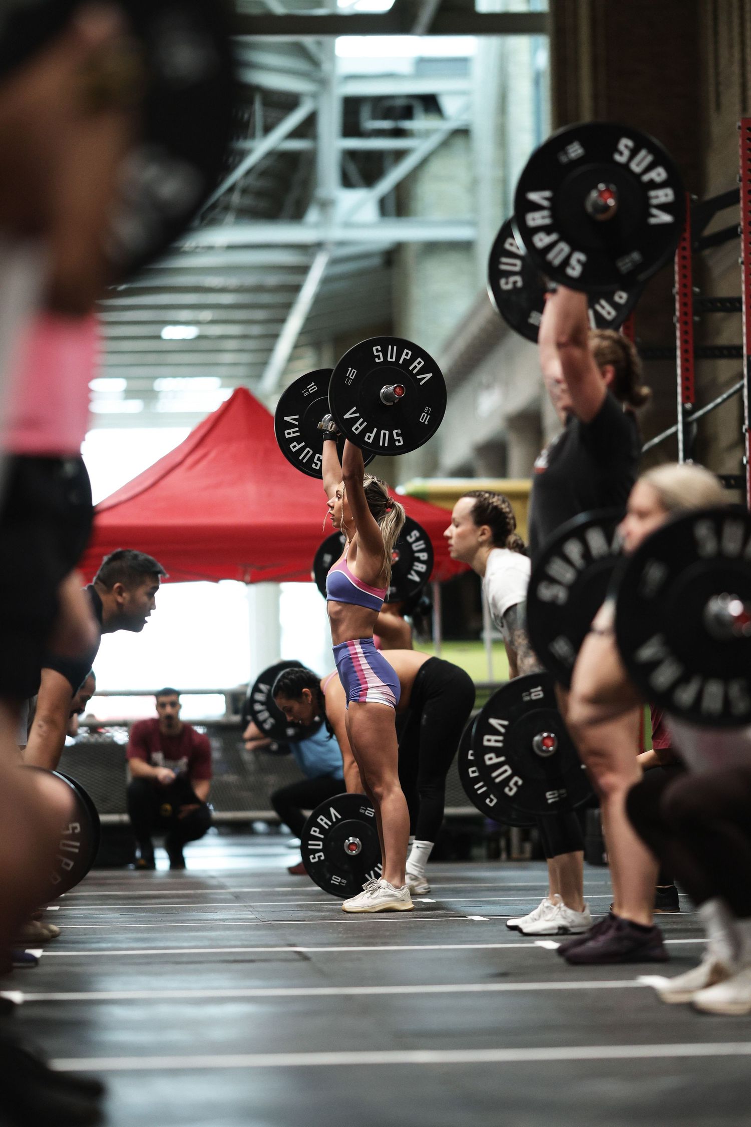 Athletes perform barbell clean exercises in sequence at a CrossFit gym with black weightlifting plates.