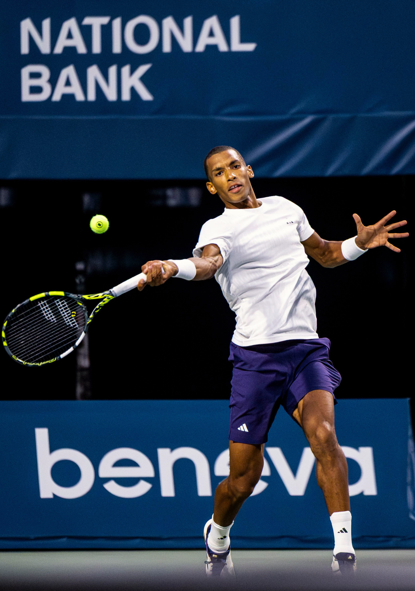 Tennis player in purple shorts and white shirt stretches to make a backhand return shot during a match.