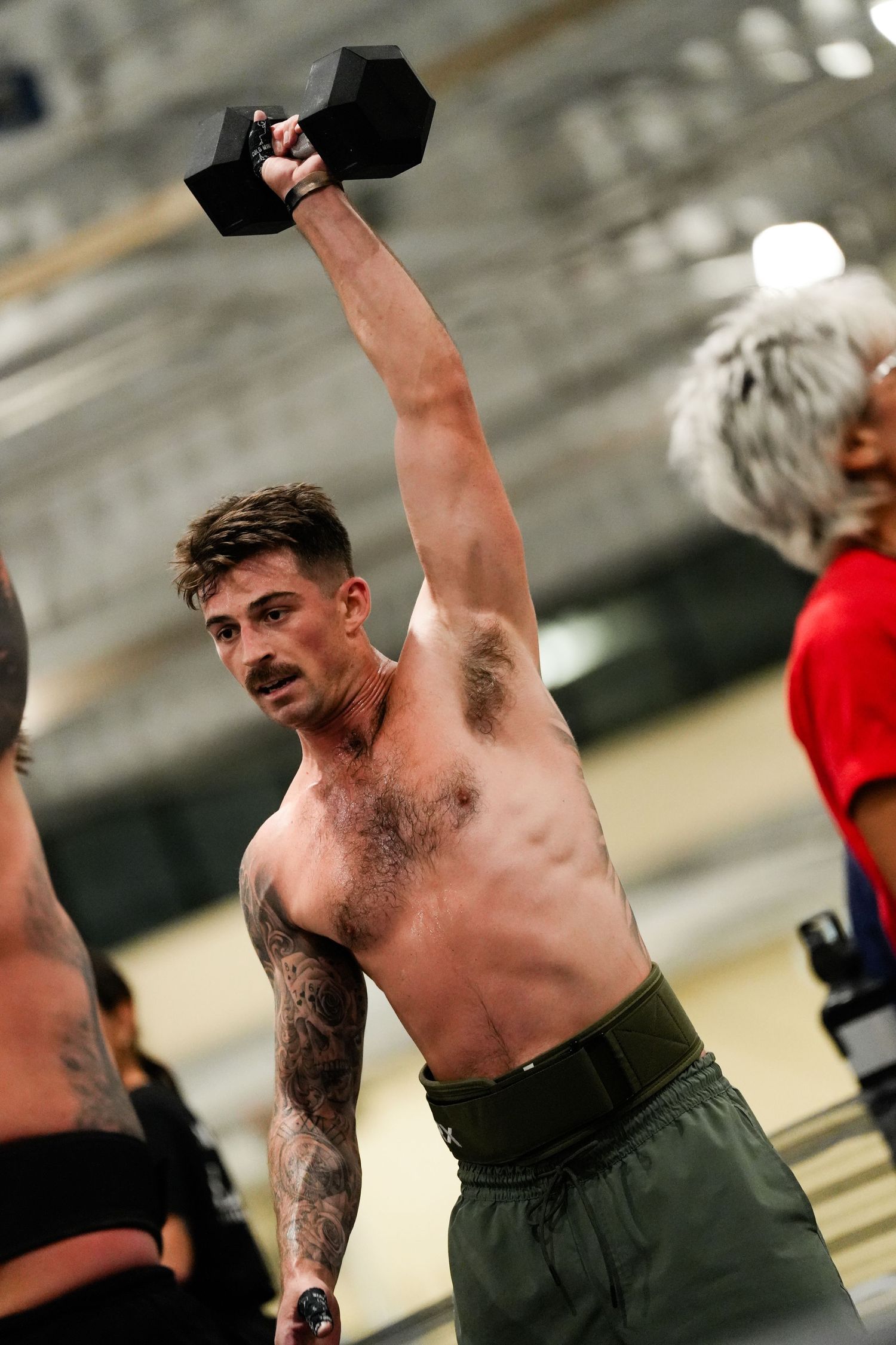Shirtless tattooed man lifting a dumbbell overhead in a gym during a CrossFit workout competition.