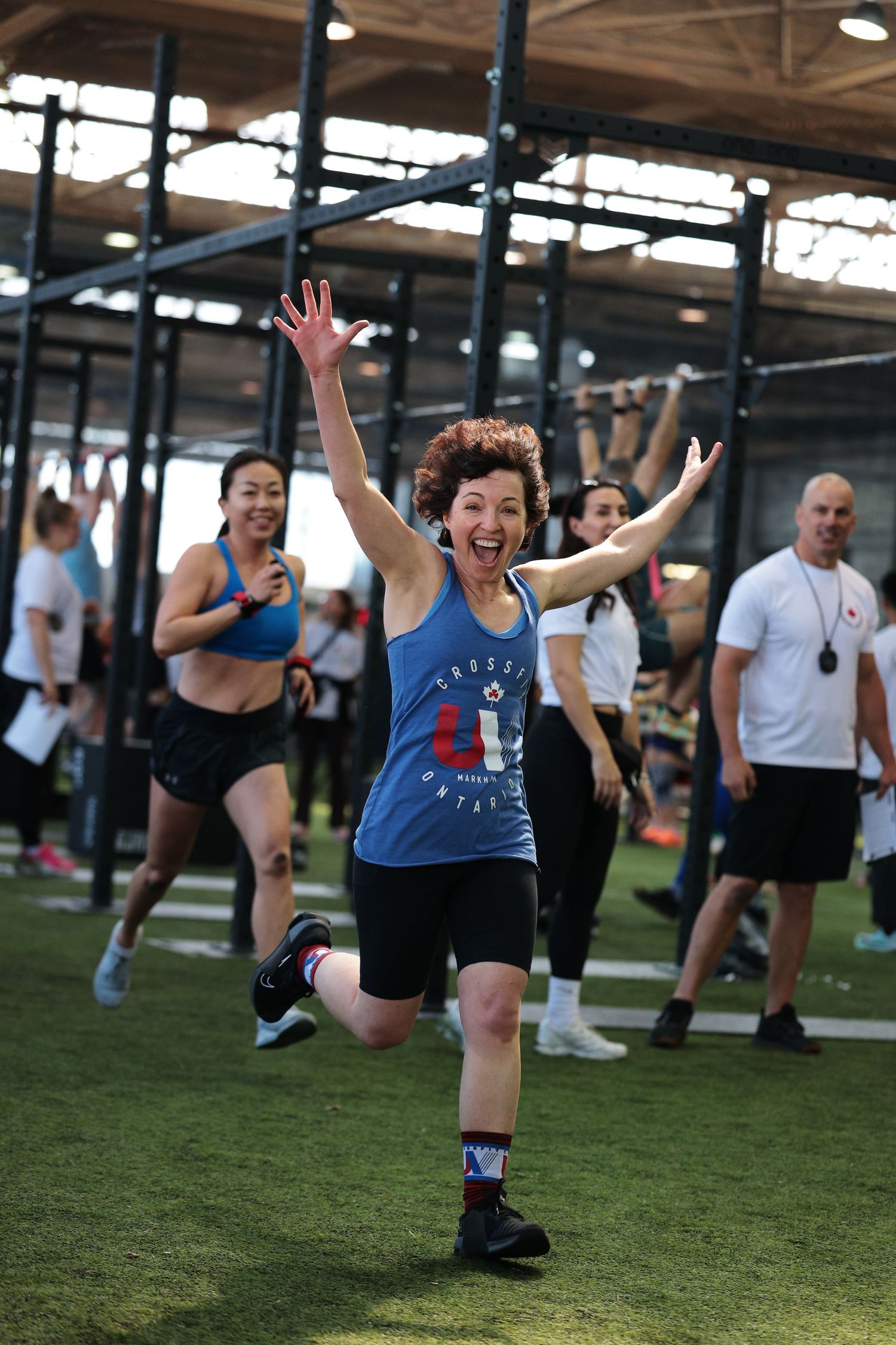 Energetic woman in blue CrossFit tank top celebrates with arms raised during group fitness workout in gym.