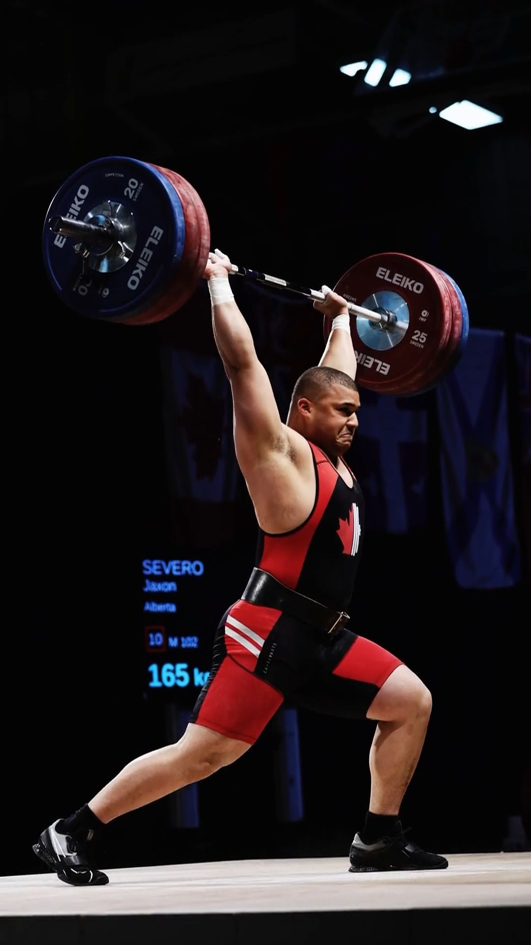 Weightlifter in red and black uniform performs a clean and jerk lift with heavy barbell during competition.