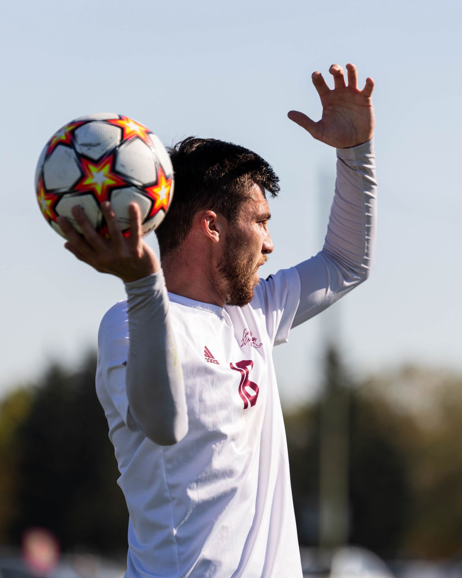 A soccer goalkeeper in a white uniform and cap reaches up to make a save during a match against the backdrop of trees.
