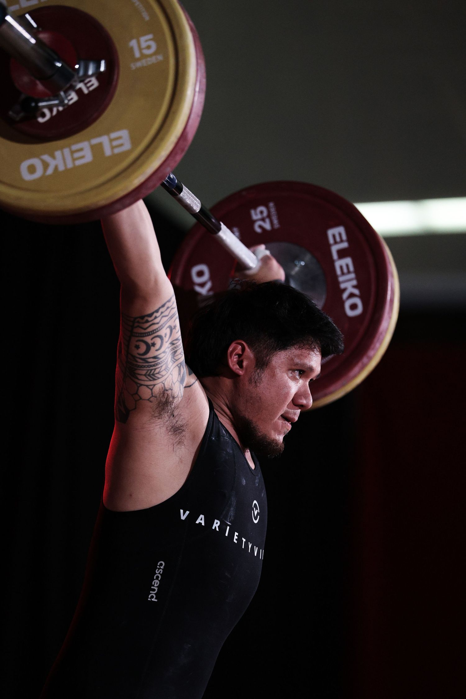 A weightlifter in a black tank top performs a clean and jerk sequence with an Olympic barbell in a dark gym setting.