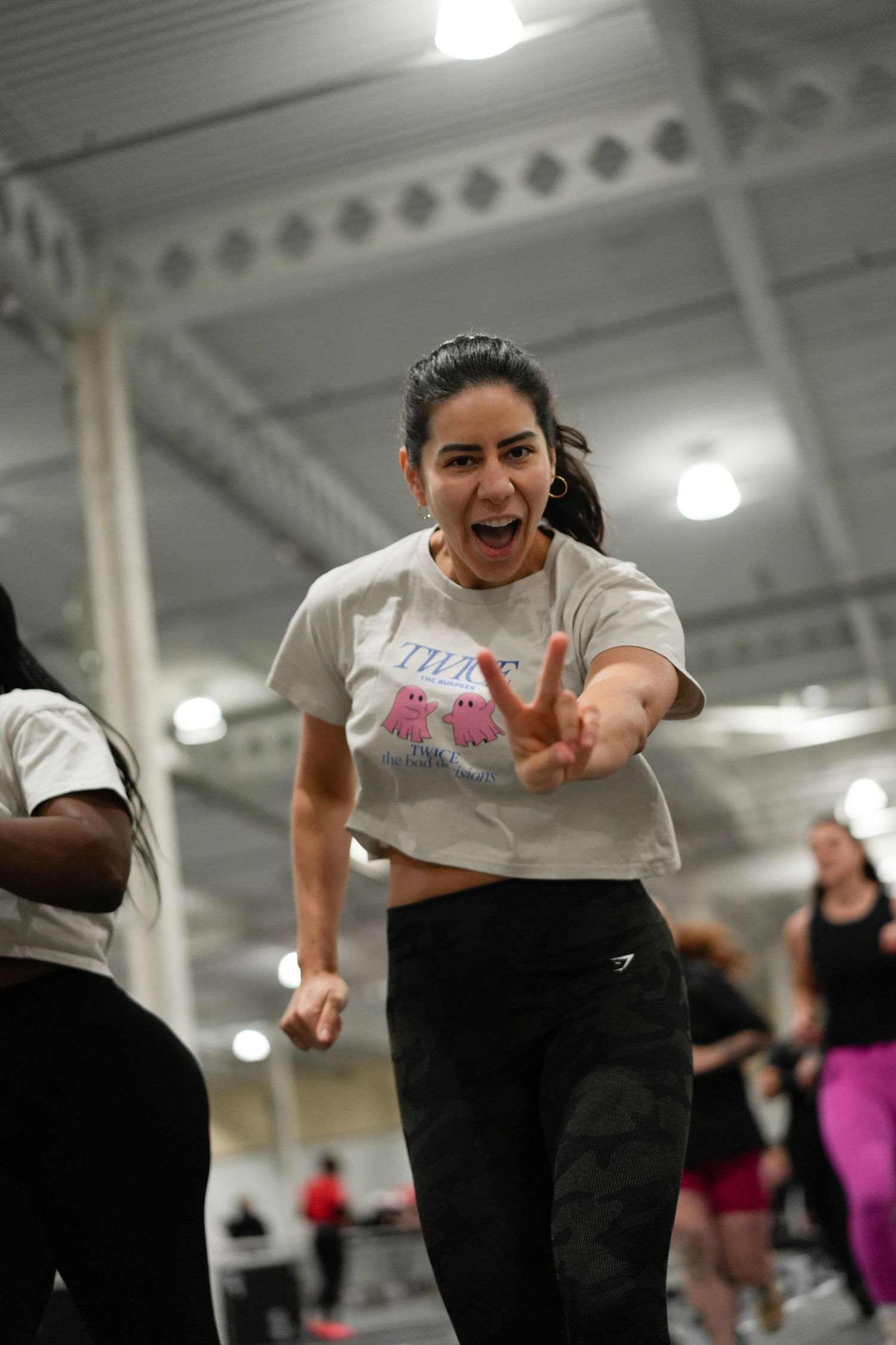Energetic woman in white crop top flashing peace sign during group fitness dance class in large gym.