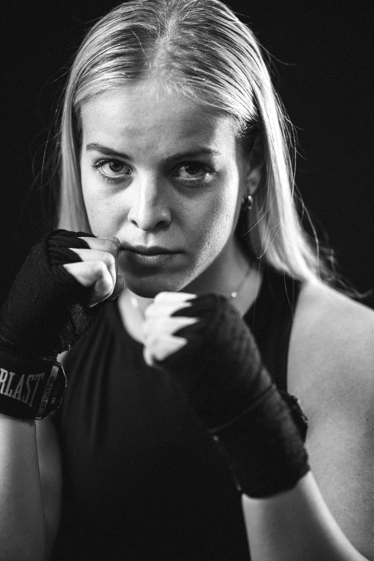 Black and white portrait of a boxer in defensive stance wearing hand wraps against dark background.