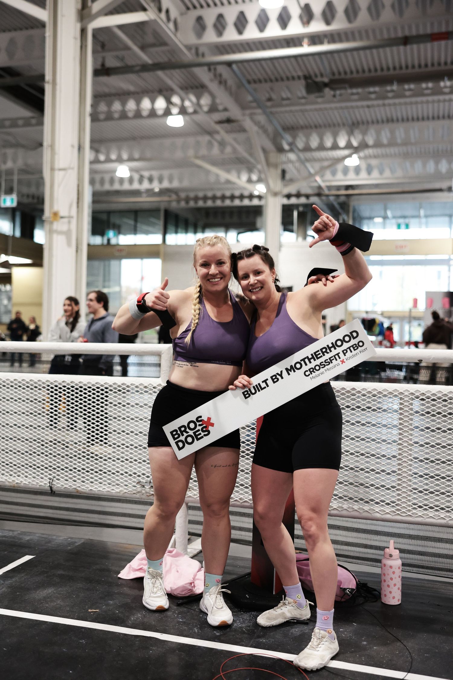 Two women pose proudly at a CrossFit competition holding a Bros & Does Built by Motherhood sign.