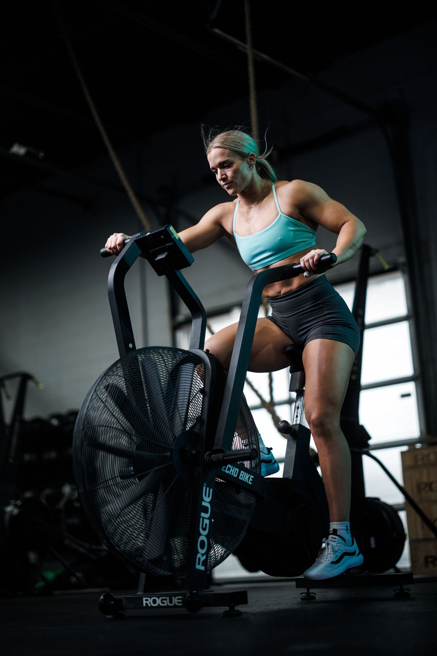 Athlete working out intensely on an air bike in a dark gym wearing turquoise tank top and navy shorts.