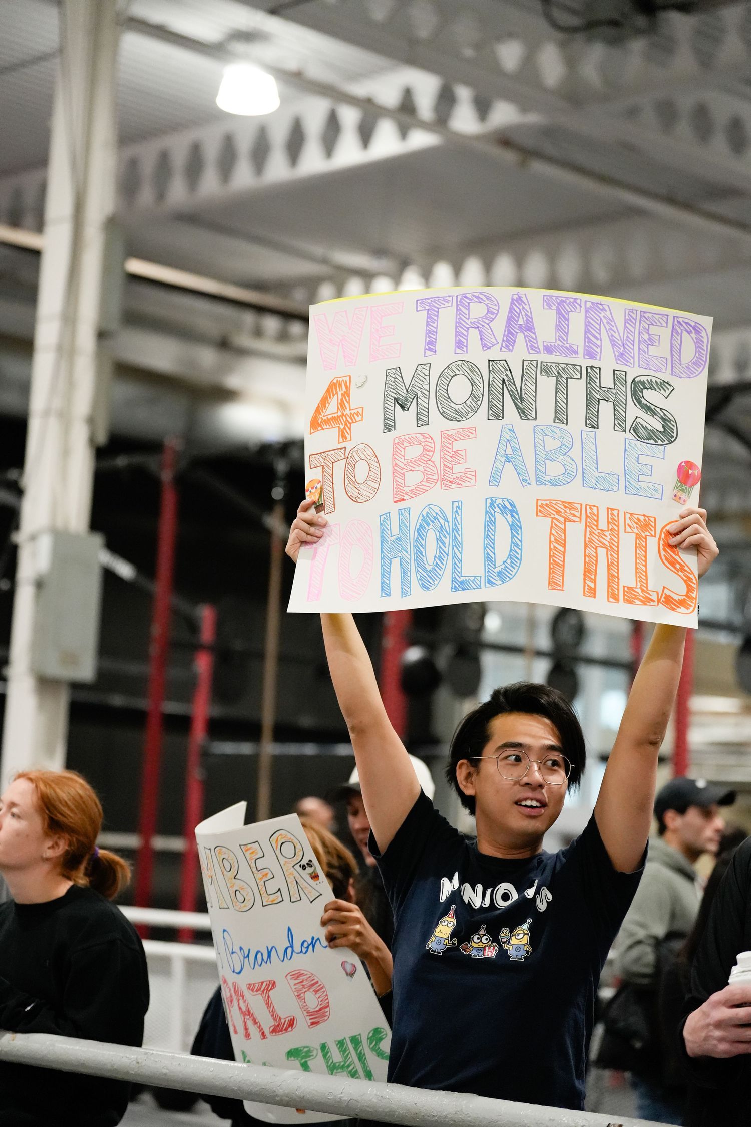Young man holds humorous sign reading 'I trained 4 months to be able to hold this' at indoor event.