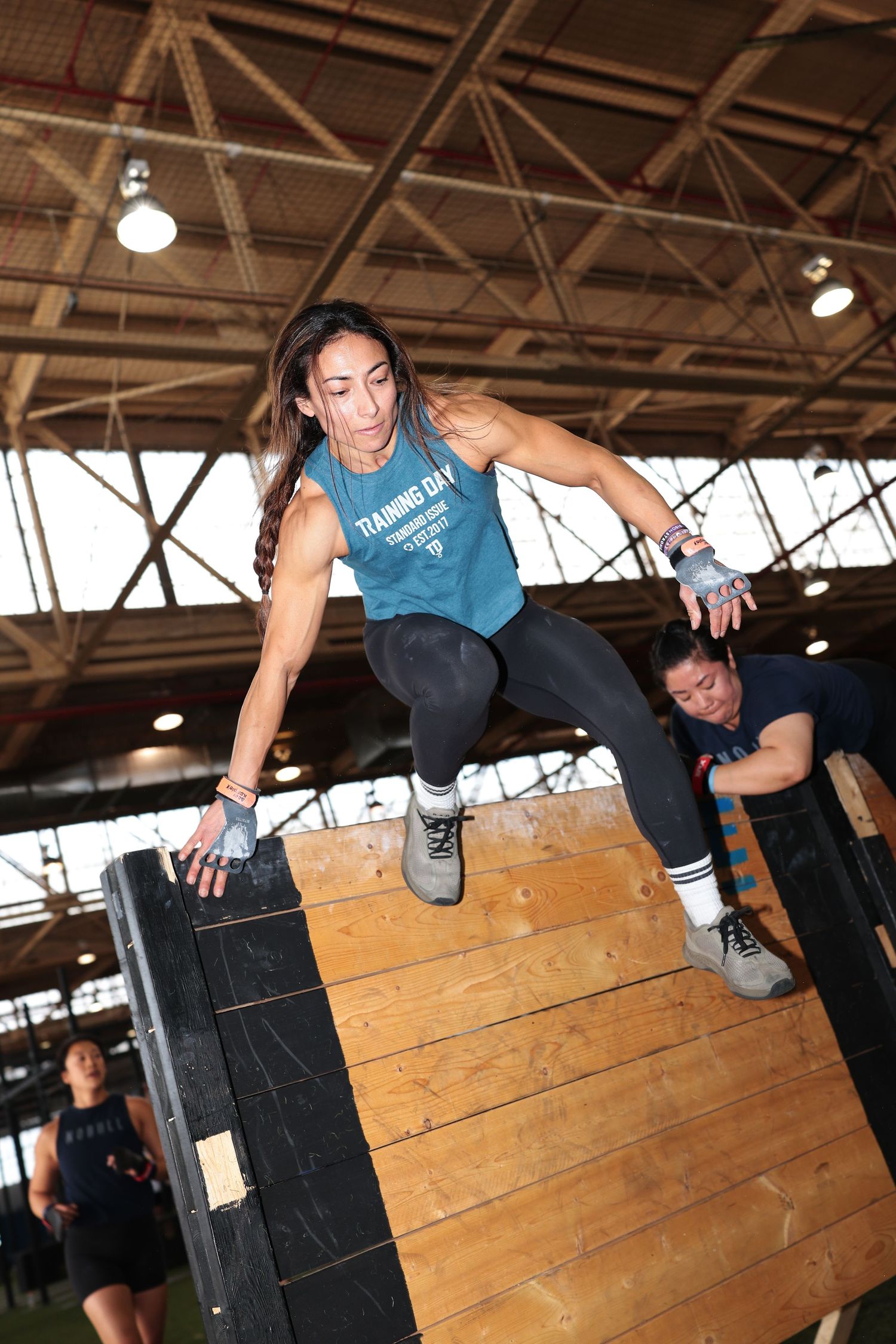Athletic woman leaping over a wooden box obstacle during an indoor fitness competition training event.