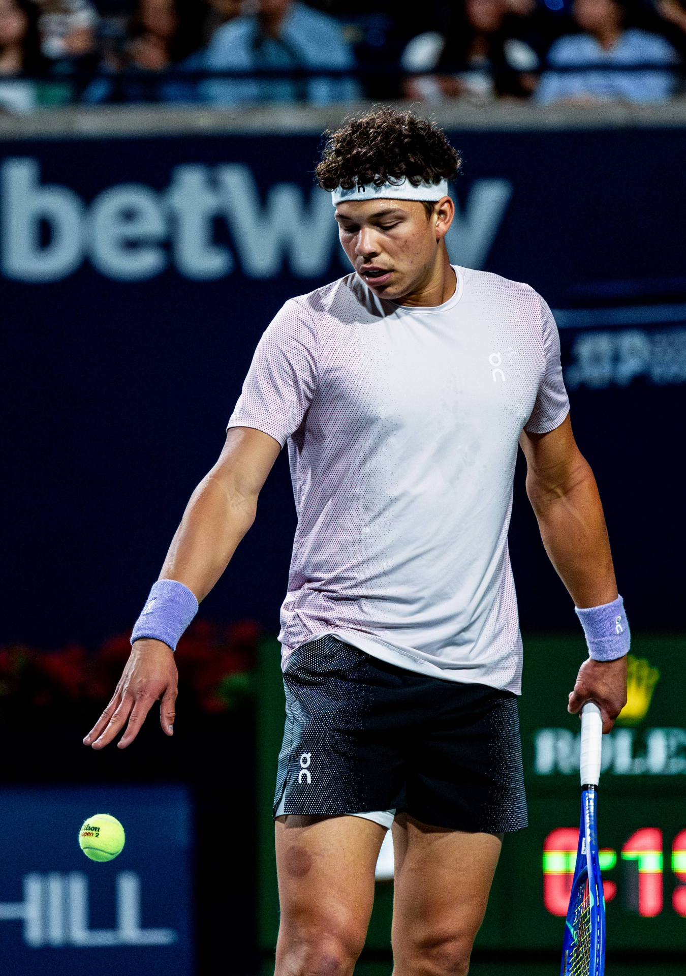 Tennis player in white shirt and black shorts preparing to serve on a professional court.