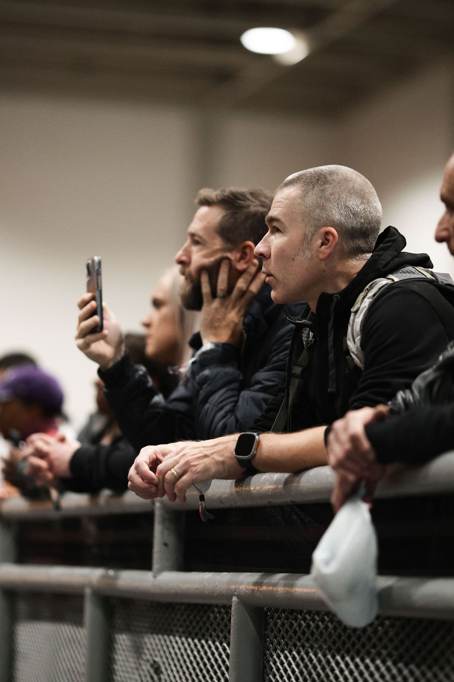 Spectators lean against a railing while watching intently and recording with phones at a sporting event.