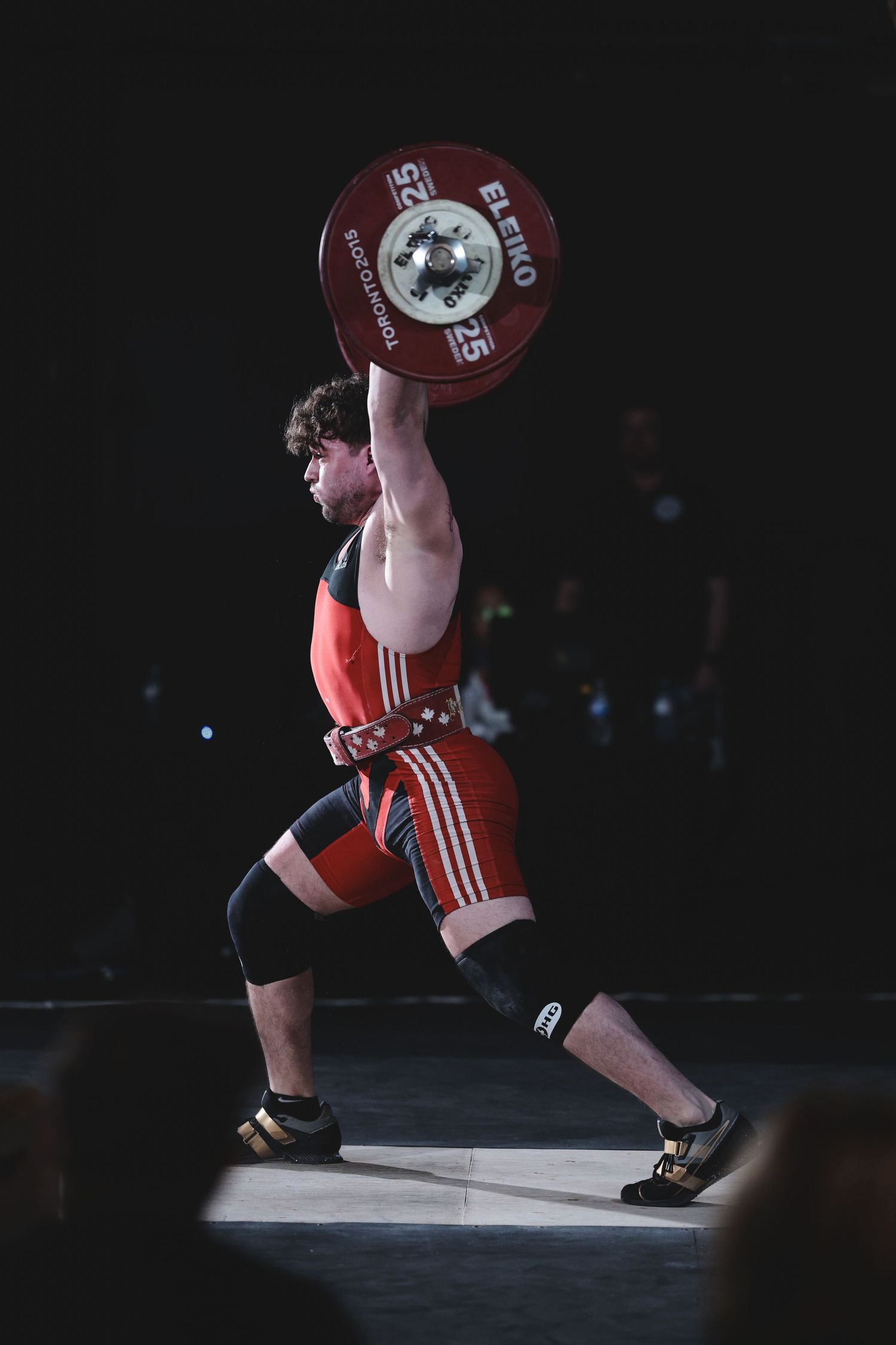 Weightlifter in red uniform performs snatch lift with heavy barbell plates during competition under spotlight.