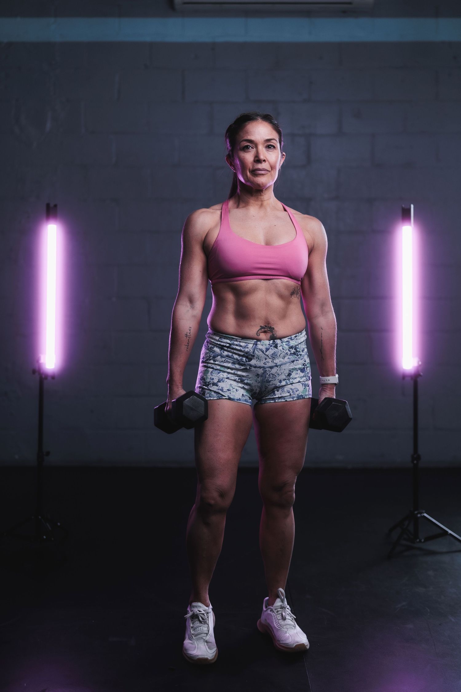 Athletic person in pink sports bra and patterned shorts holds dumbbells while posing between purple LED lights in dark gym.