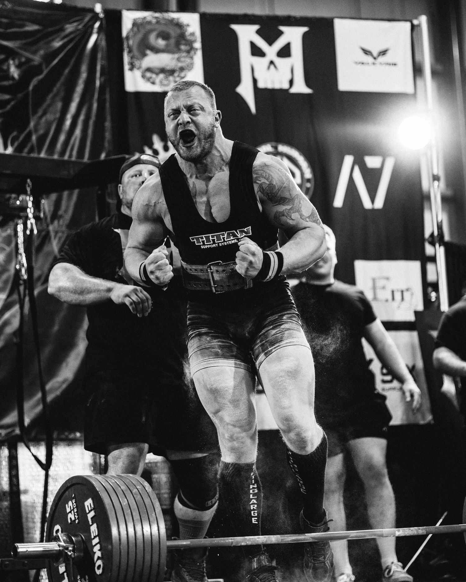 Three powerlifters perform deadlifts with heavy weights in a black and white competition setting.