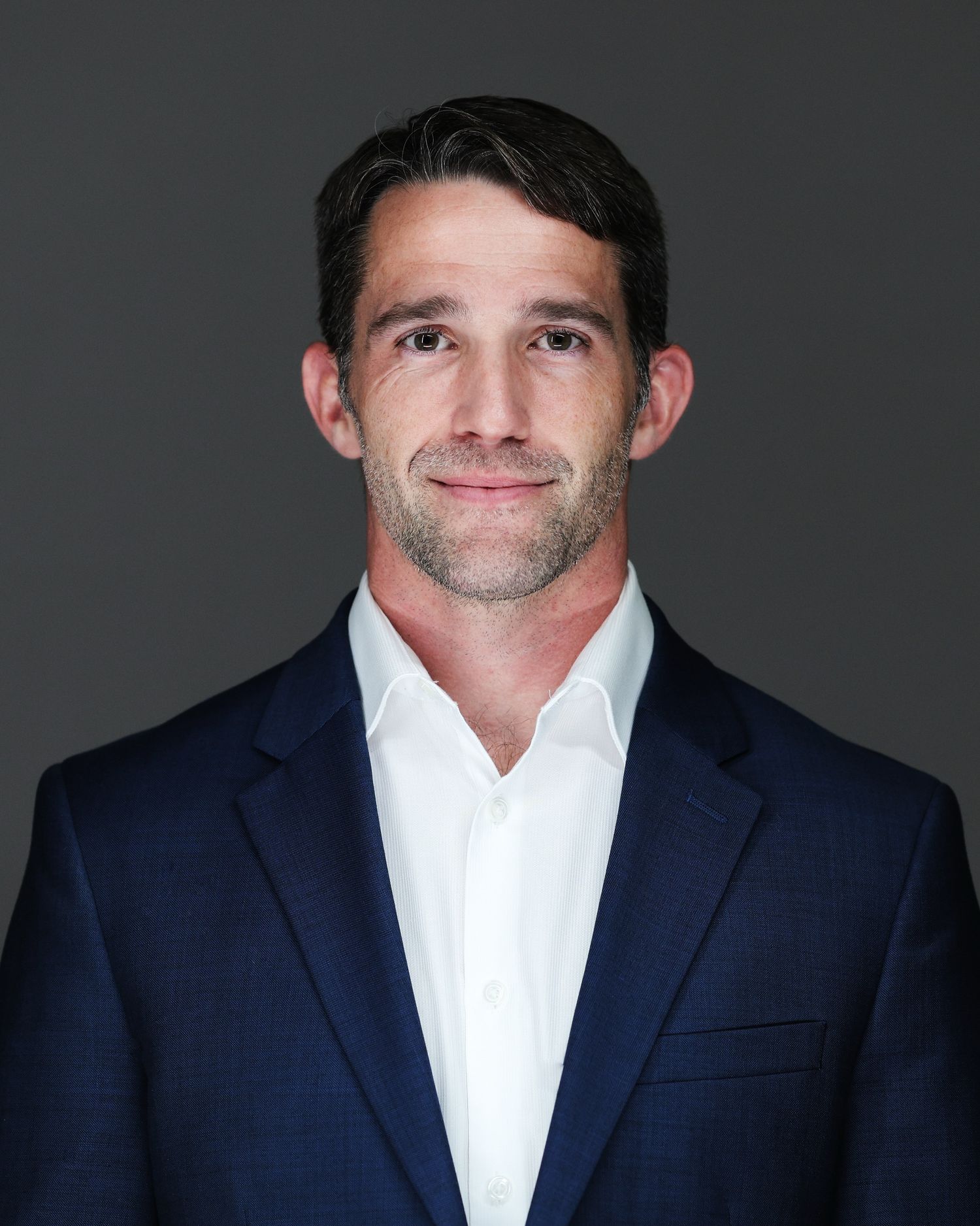 Professional headshot of a person in a navy suit and white dress shirt against a dark gray background.