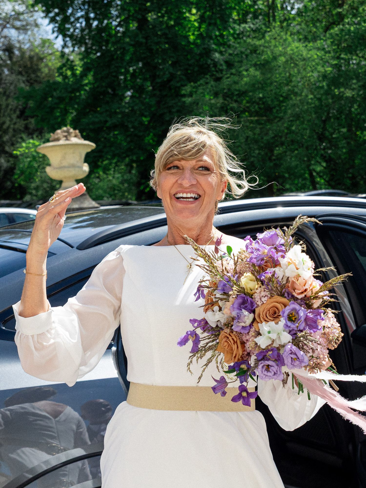 A joyful person in a white dress holds a vibrant purple and peach floral bouquet while celebrating in front of a car.