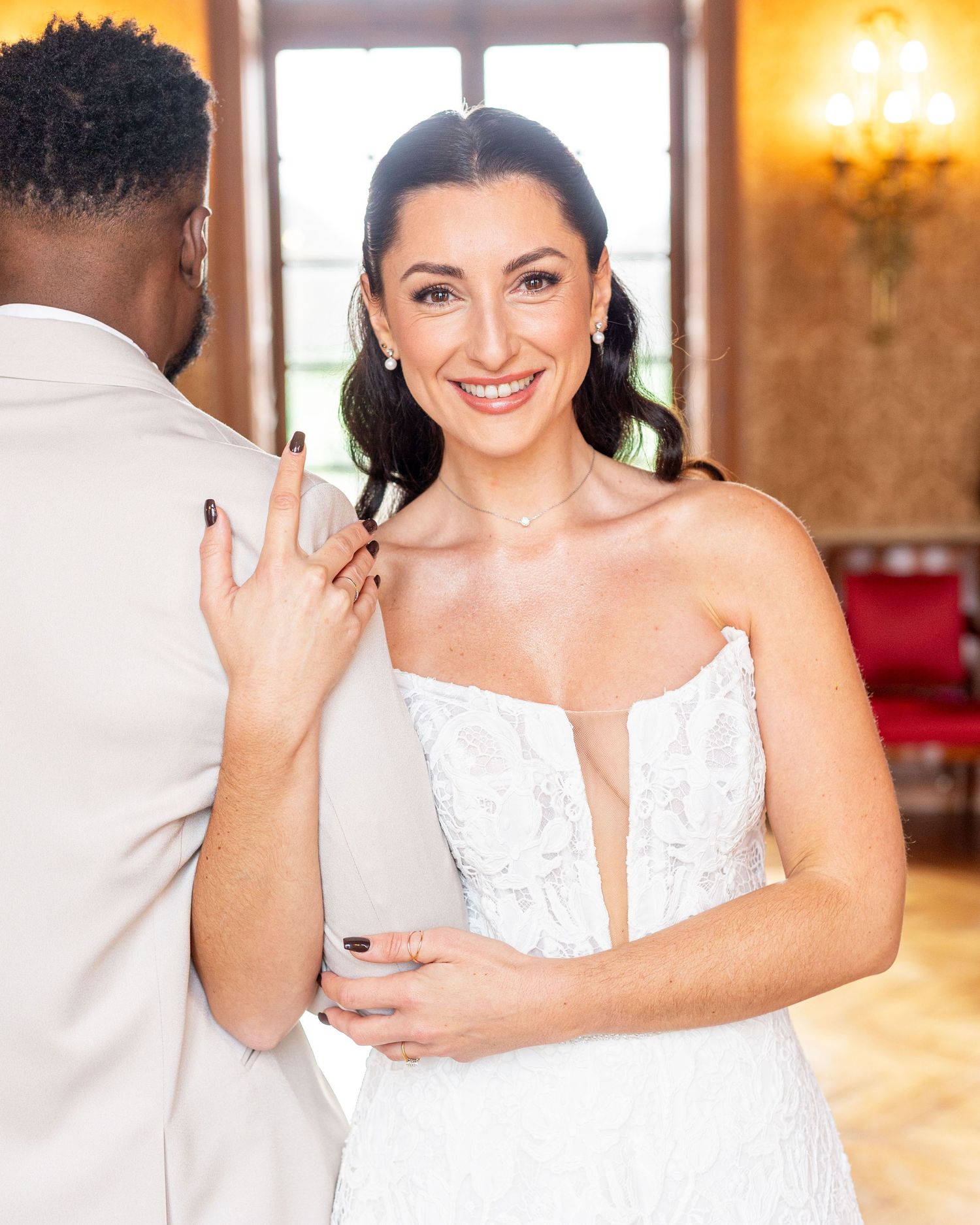 Bride in white lace gown smiles brightly in sunlit ballroom setting.