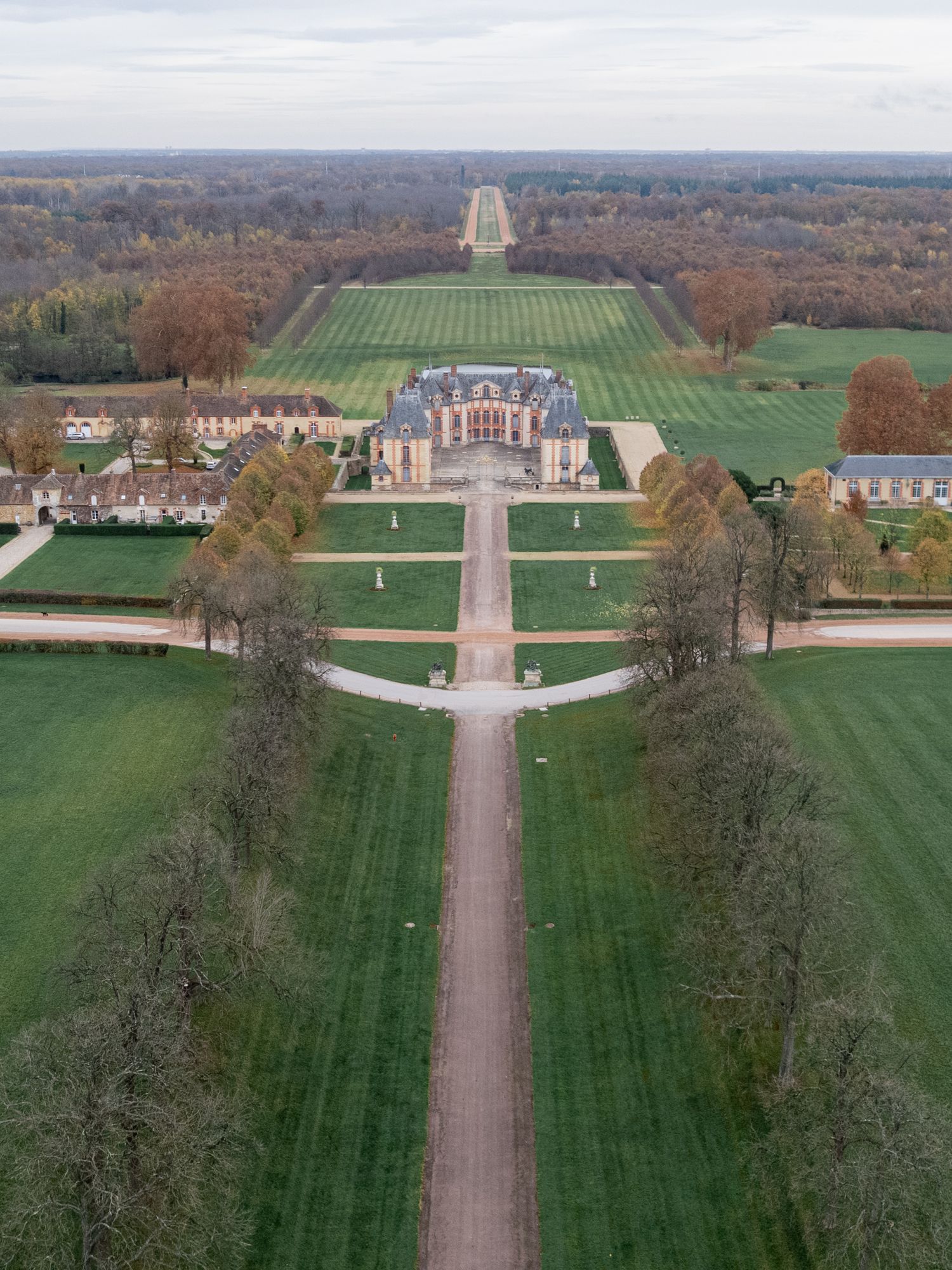 Aerial view of a grand estate with manicured gardens, tree-lined paths, and expansive green lawns stretching to the horizon.